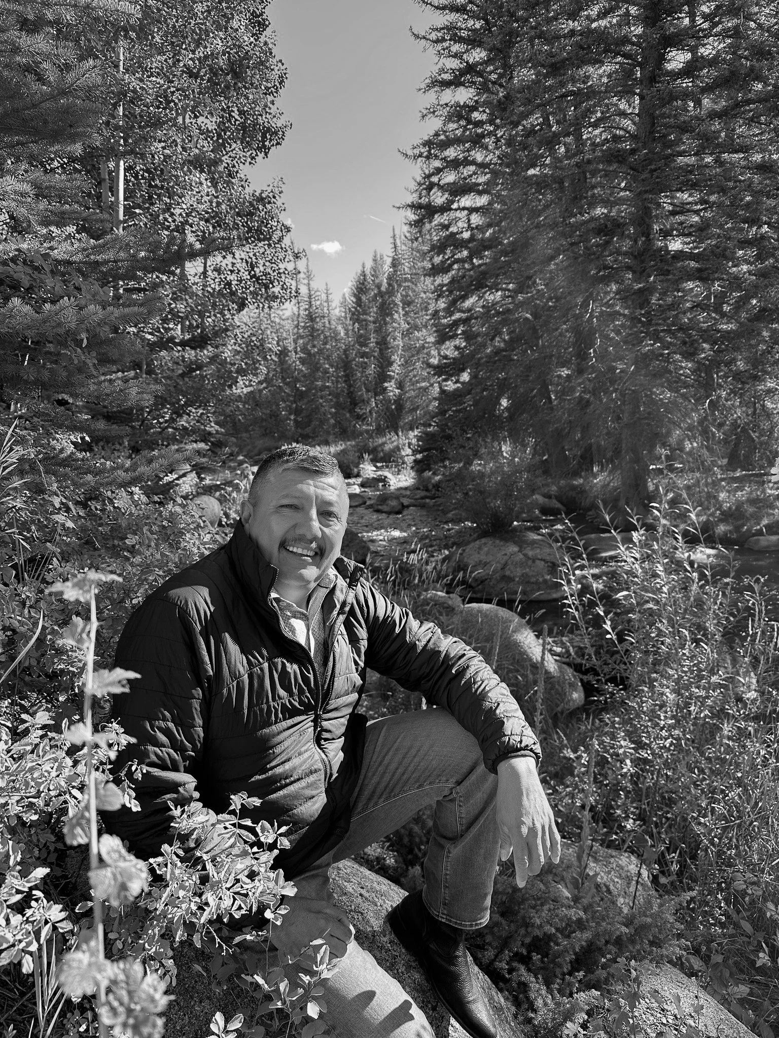 A man smiling and sitting on a rock near a stream in a forest with tall pine trees, some rocks, and bushes, in black and white.