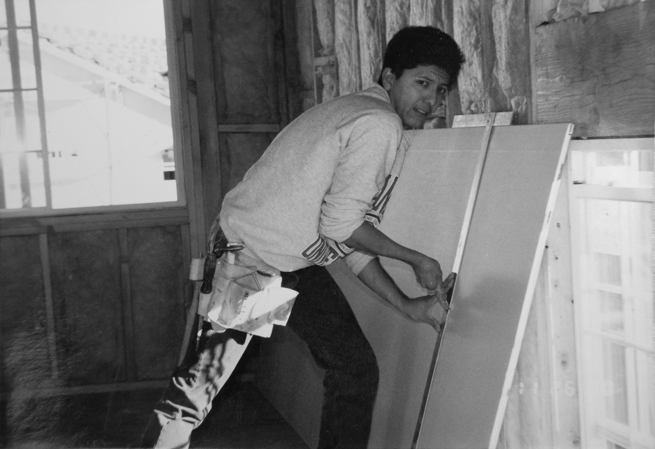 A man in a long-sleeve shirt, with paint tools hanging from his pocket, is painting a wall or large panel inside a wooden structure. He is smiling and looking at the camera while working.