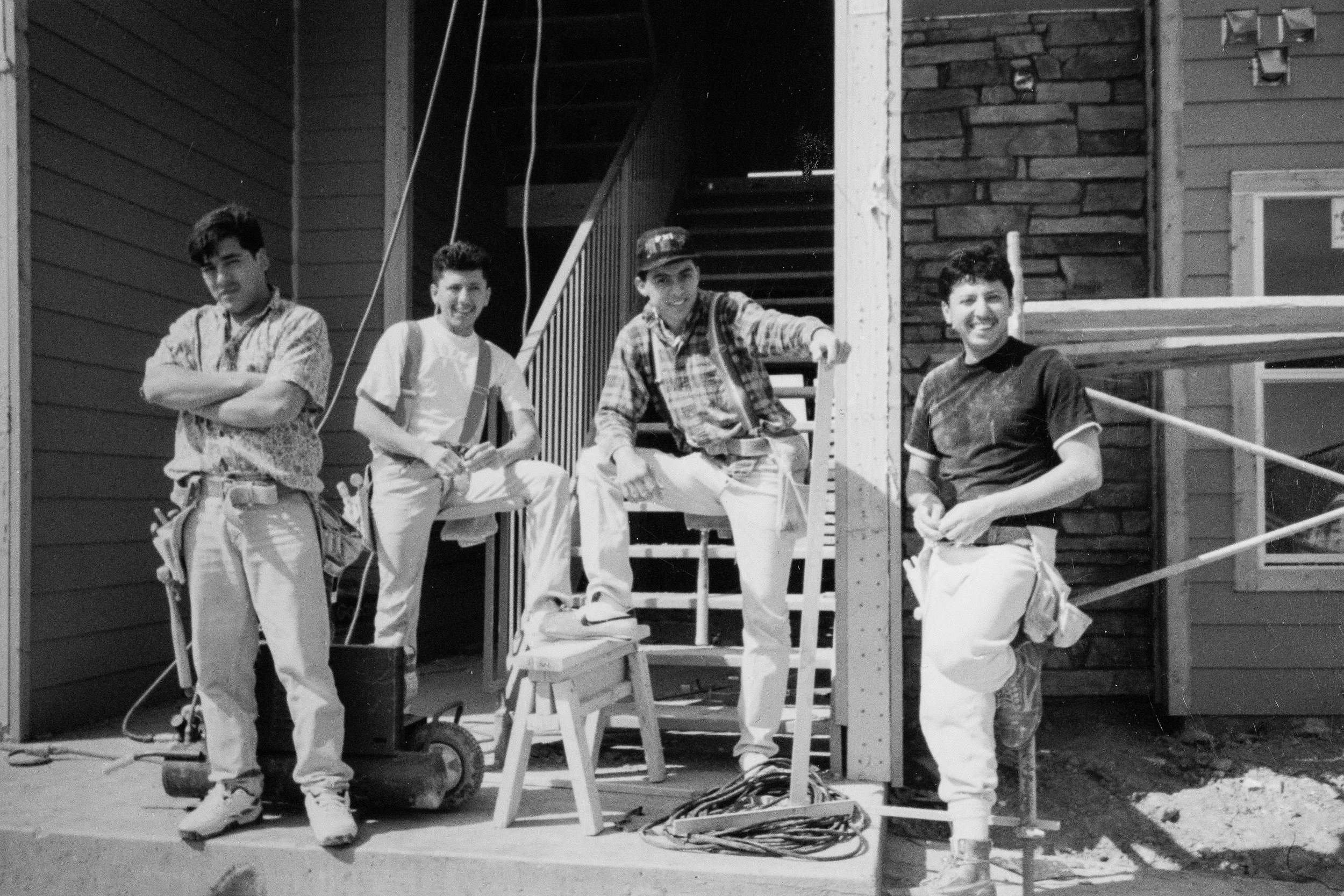 Black and white photo of four young men working on a construction site, standing and sitting near a partially built wooden staircase and house.