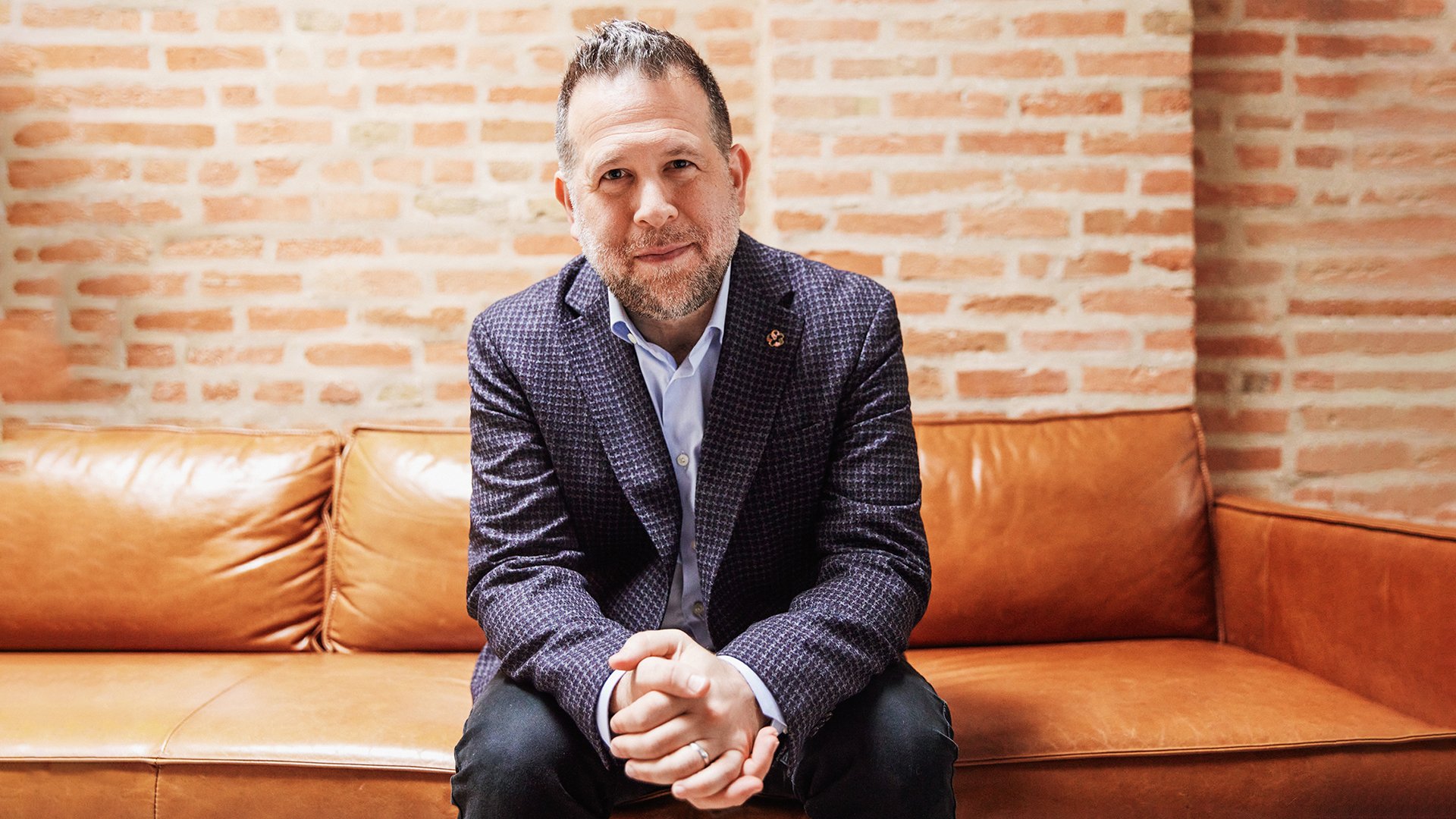 Portrait of Matthew Jacobson seated on a leather sofa in a brick-walled studio, wearing a patterned blazer