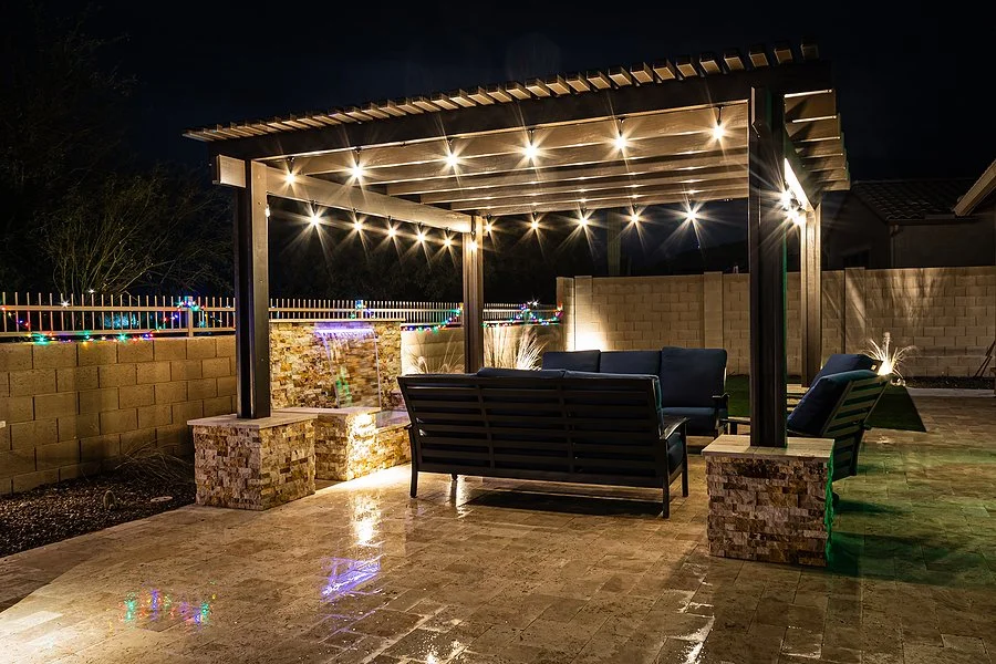 Nighttime outdoor patio with a wooden pergola decorated with string lights, stone base, and outdoor furniture, including sofas and chairs, on a wet tiled surface with a brick wall and holiday lights in the background.