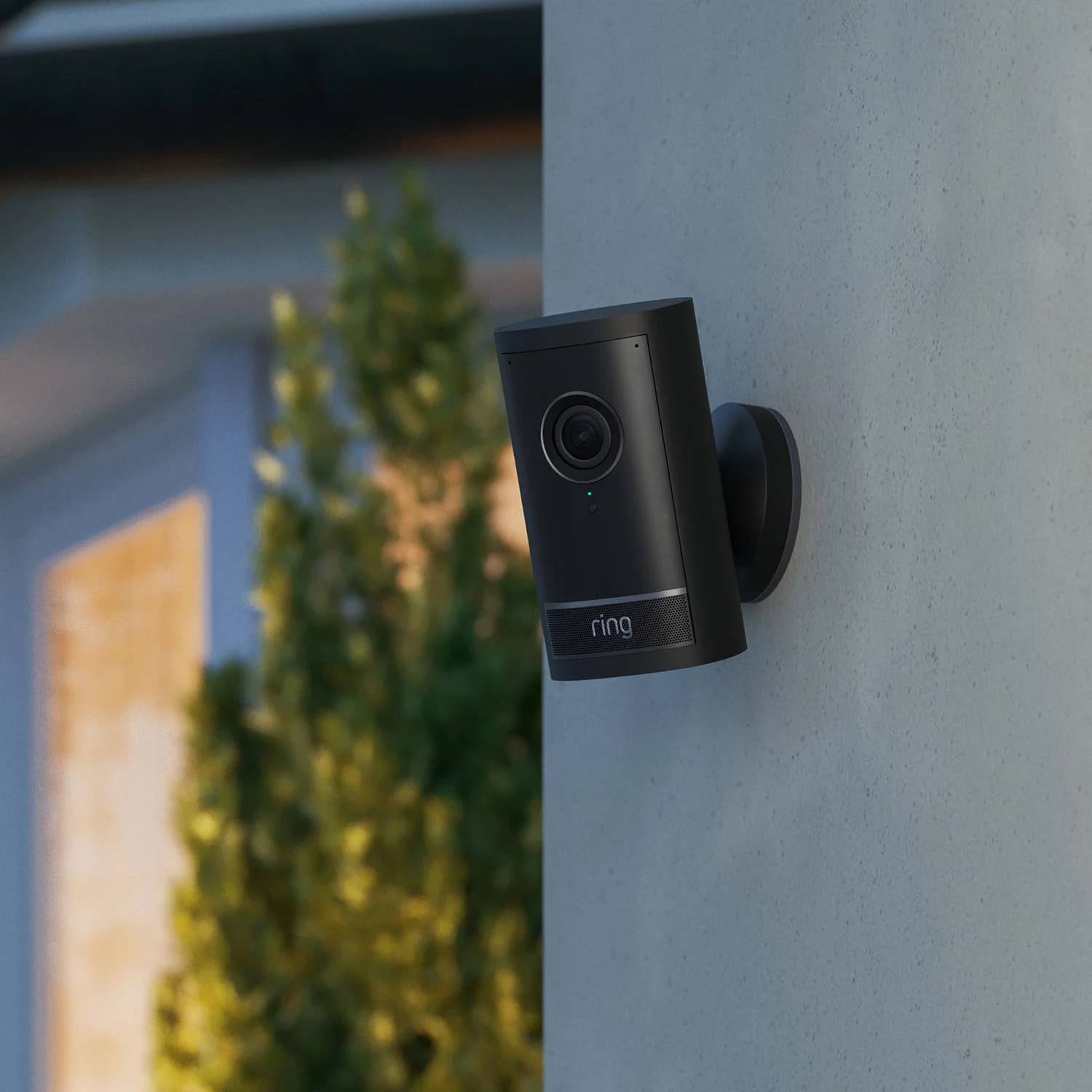 A black Ring security camera mounted on a light-colored outdoor wall with trees and a house in the background.