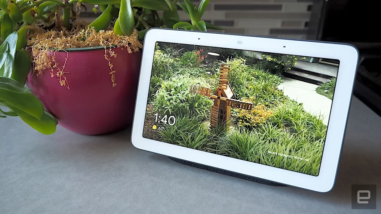 A digital picture frame displaying a garden scene with a small wooden windmill among green plants and trees, placed next to a pink pot with a succulent plant on a light gray surface.