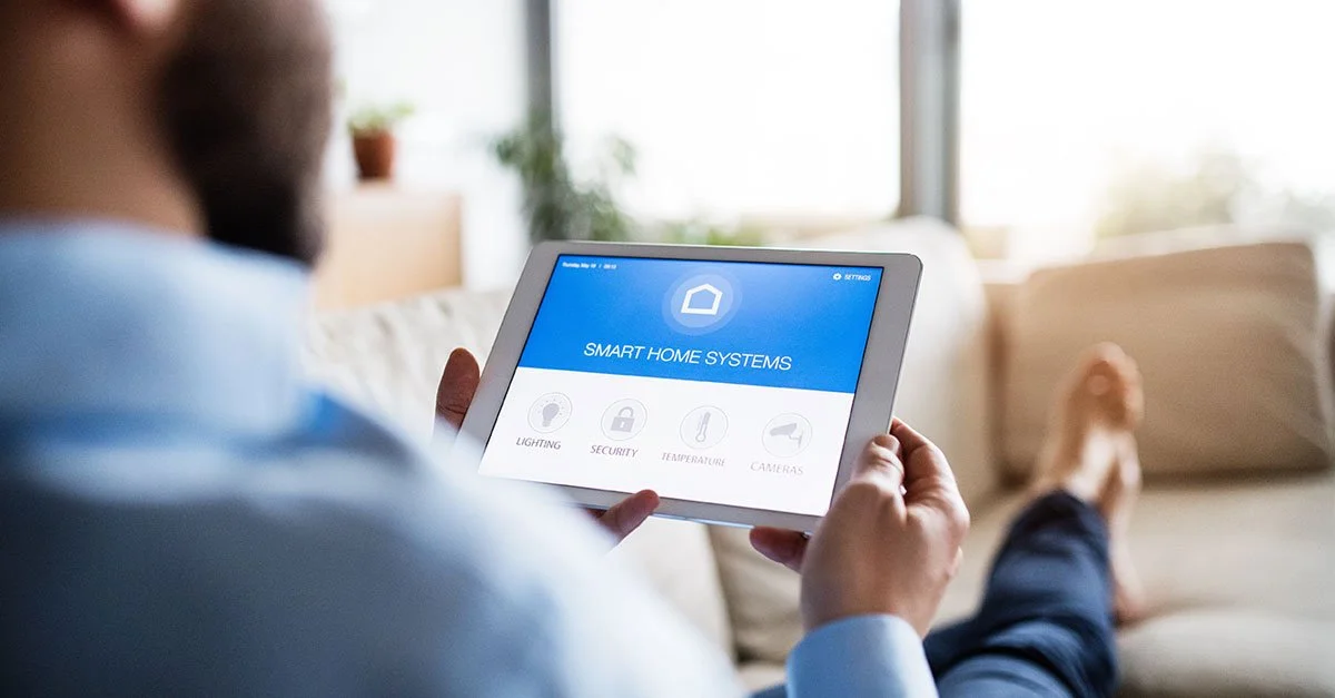 Person holding a tablet with a smart home system app, sitting on a beige sofa in a well-lit living room.