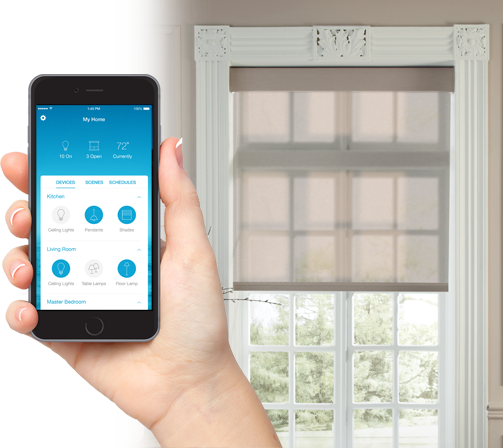 Person holding a smartphone with a smart home app open, in front of a window with a white curtain and molding in a room.