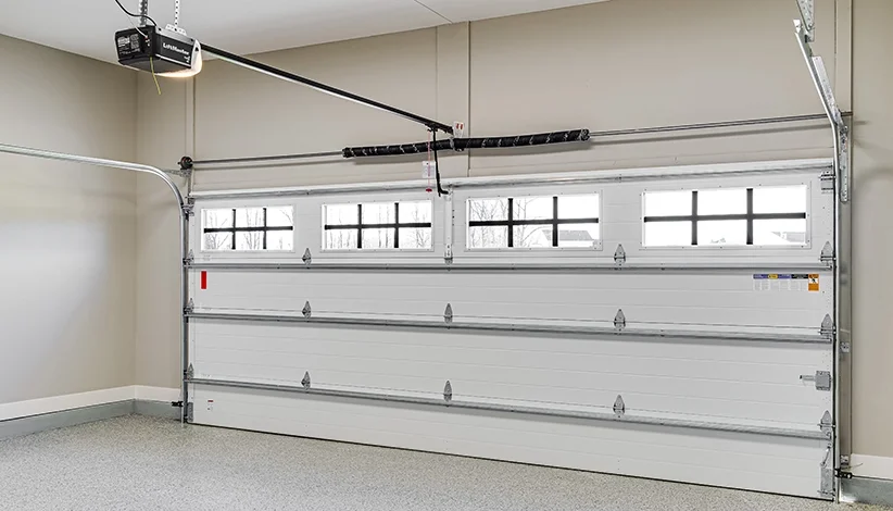 A closed white garage door with four windows near the top, installed in an empty garage with beige walls and a speckled gray floor.