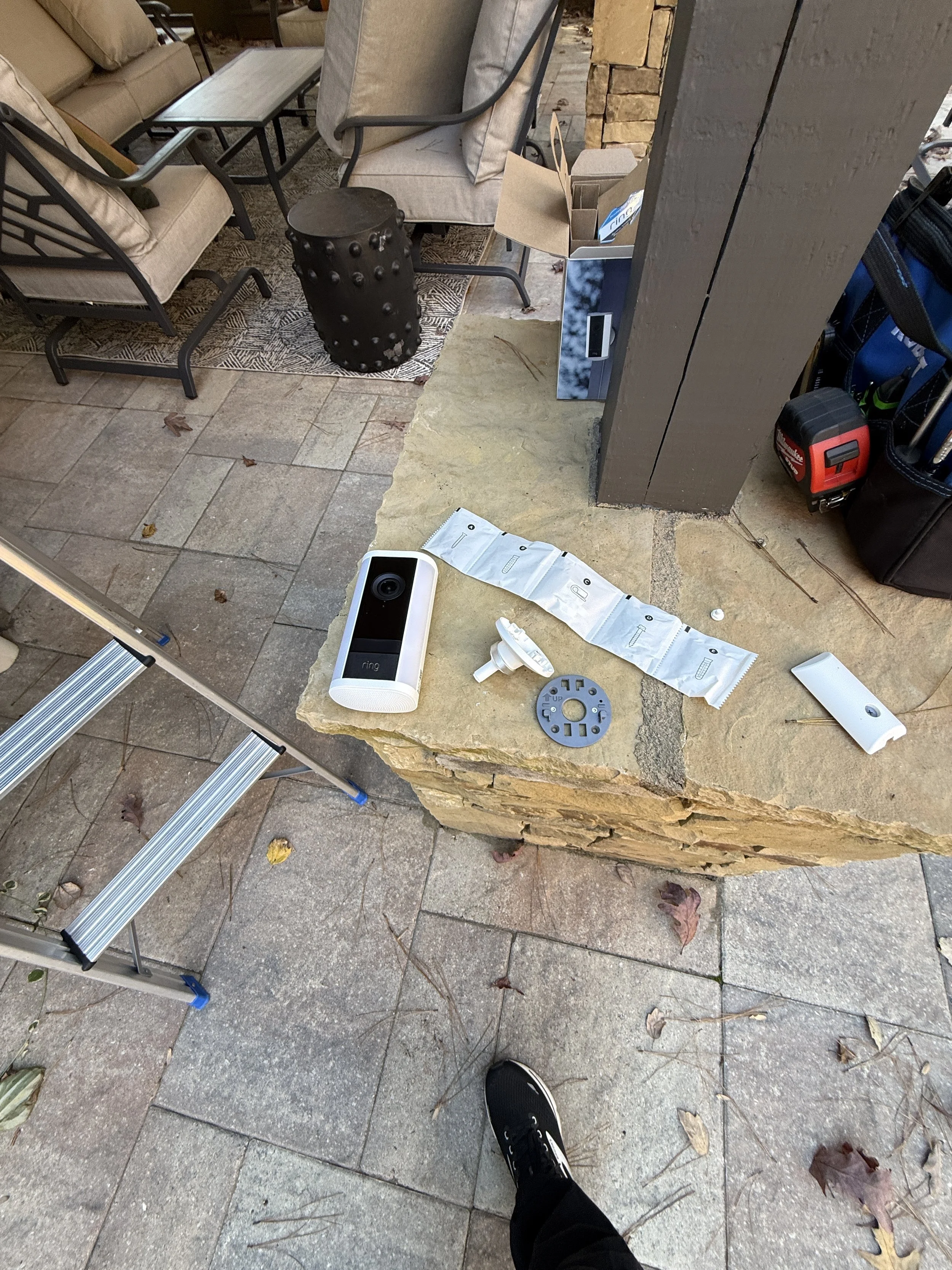 Outdoor patio with chairs, table, and various disassembled electronic devices and tools placed on a stone ledge, with a person's foot visible in the foreground.