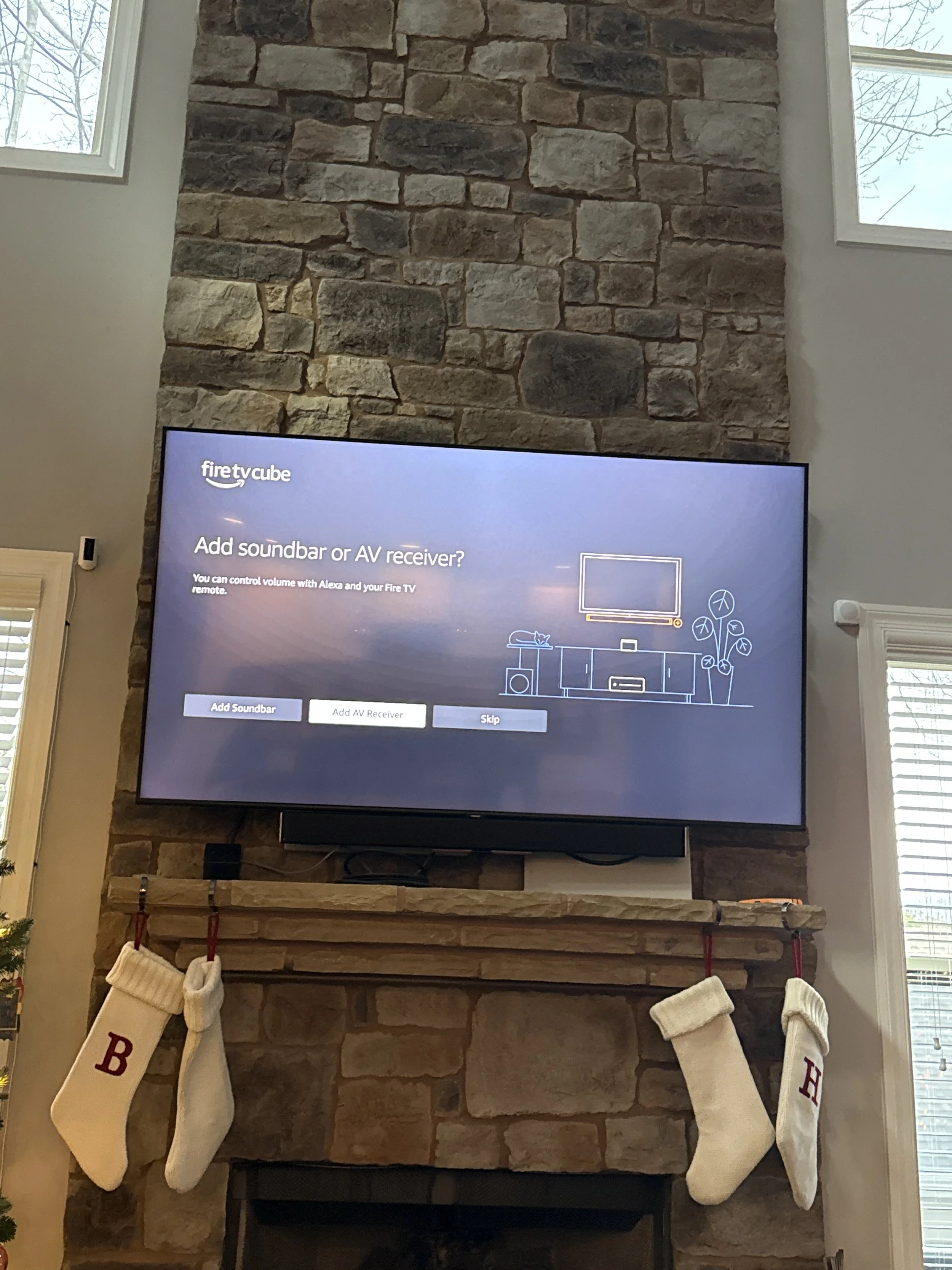 TV screen displaying setup options for adding soundbar or AV receiver, mounted above a fireplace with stockings hanging on each side, within a living room with stone wall and window.
