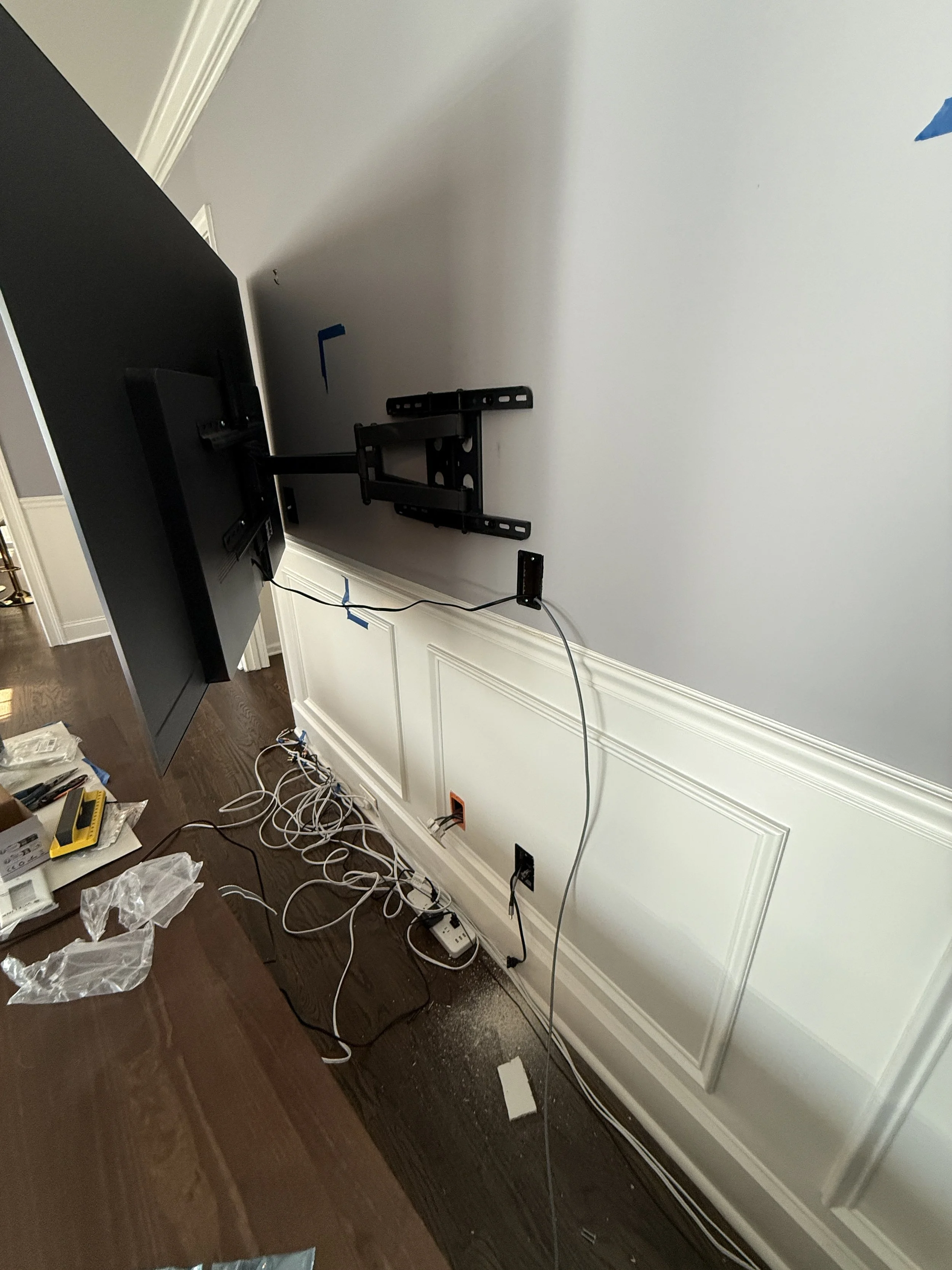 TV wall mount partially installed with wires hanging down, in a room with hardwood flooring and white wainscoting.
