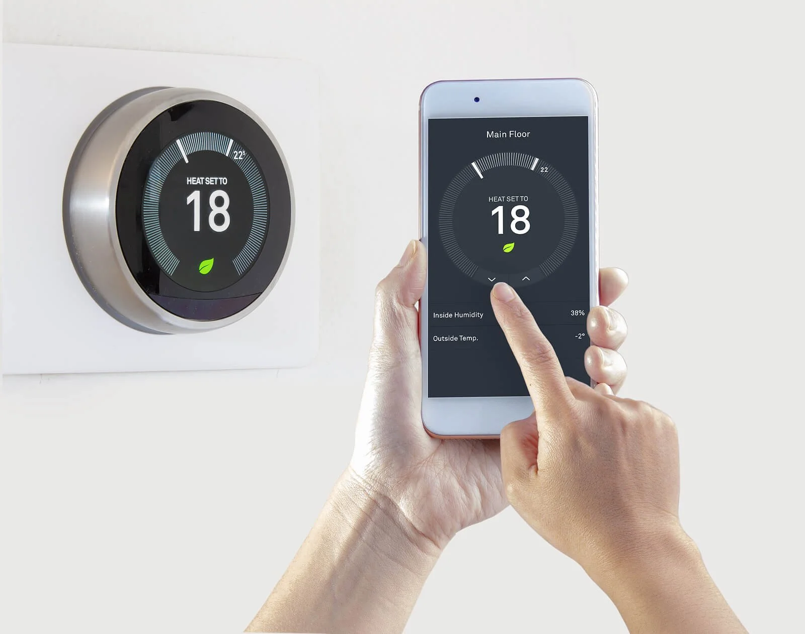 A person controls a smart thermostat using a smartphone app, adjusting the temperature set to 18 degrees. The thermostat display shows the same temperature with a green leaf icon, indicating eco mode. The app shows indoor humidity at 38% and outdoor 
