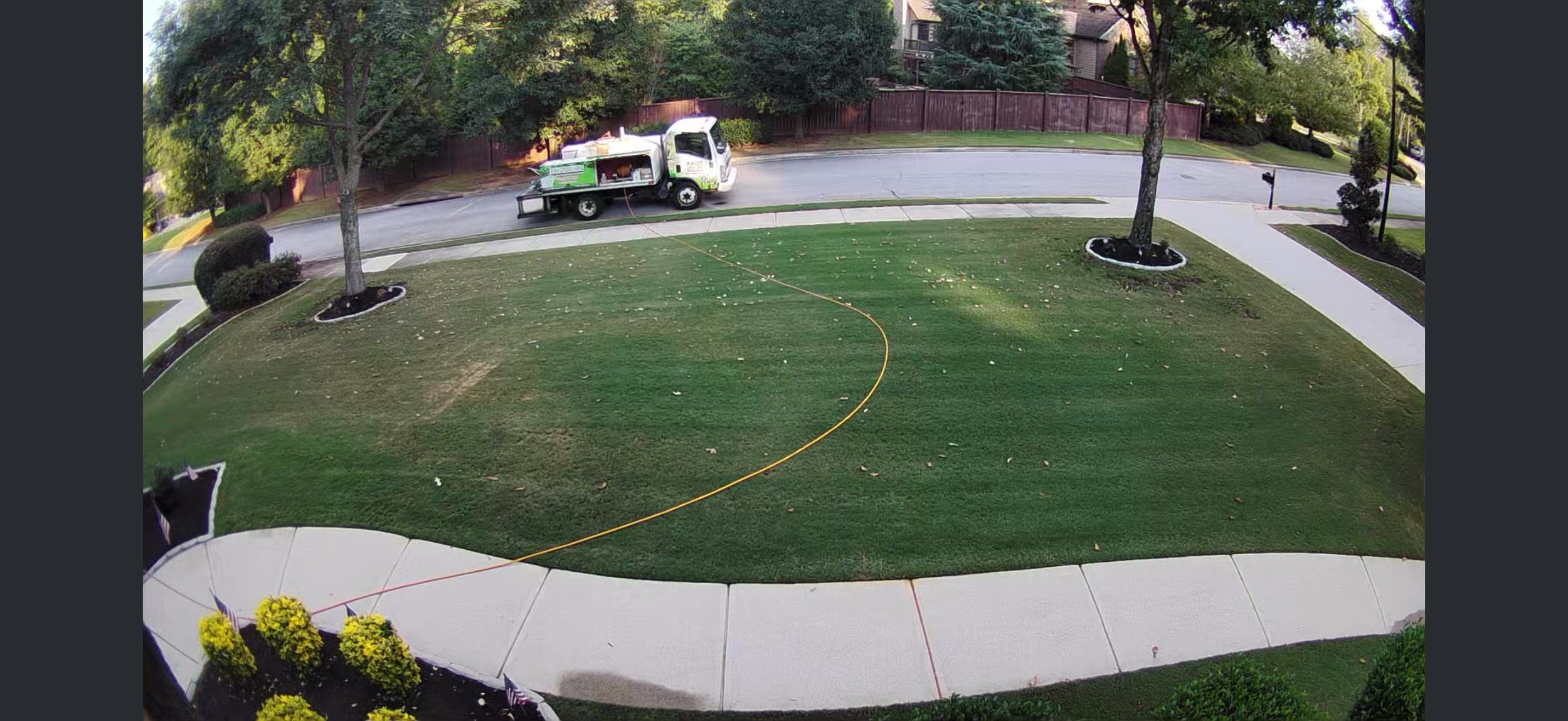 A lawn with a yellow garden hose stretching across it, connected to a nearby hose reel in the front yard. Two trees with mulch beds are present, along with a sidewalk and street. A white utility truck is parked on the street.