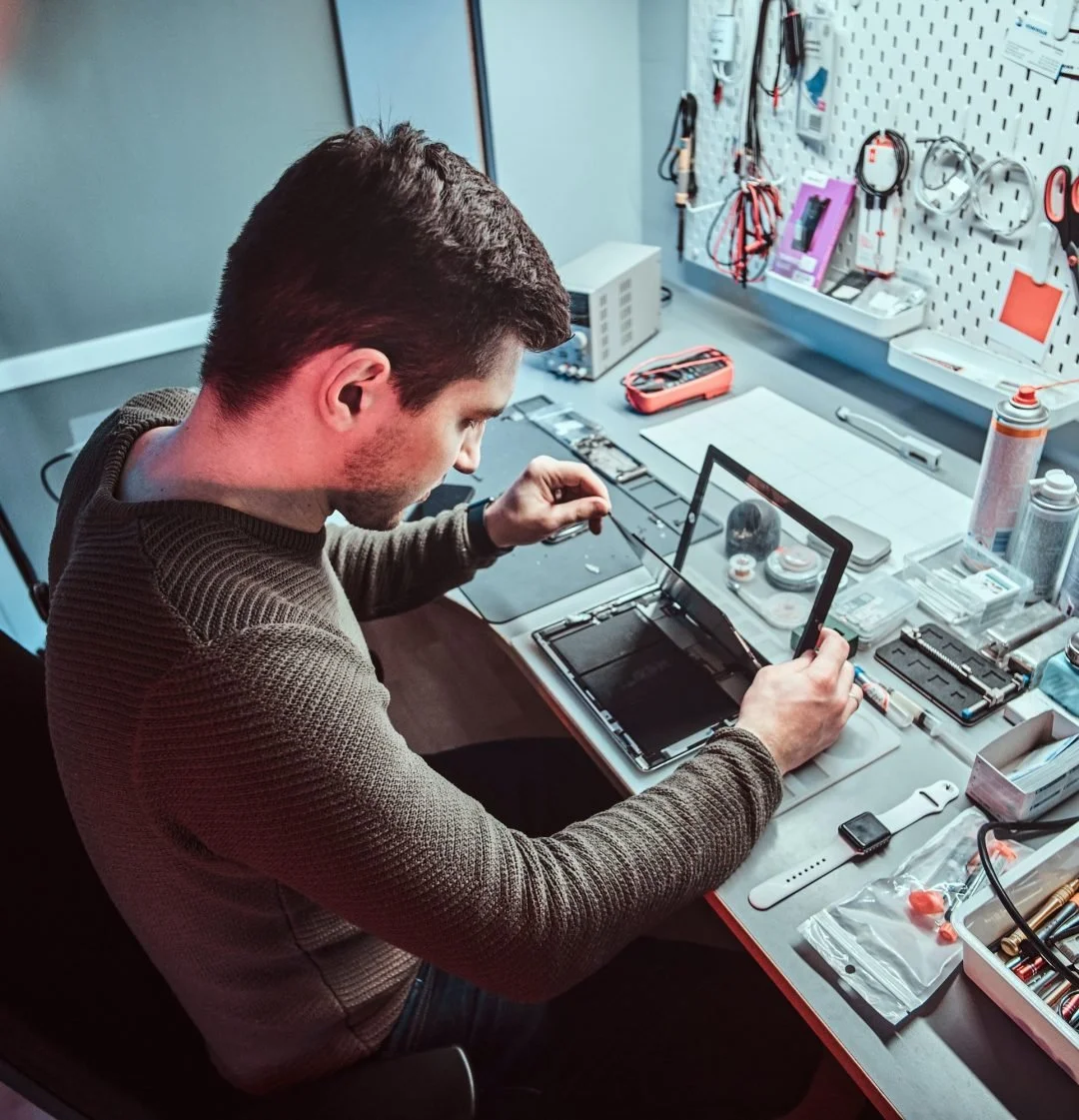 A man working in an electronics repair workshop, examining a disassembled electronic device with tools and equipment on the workbench.