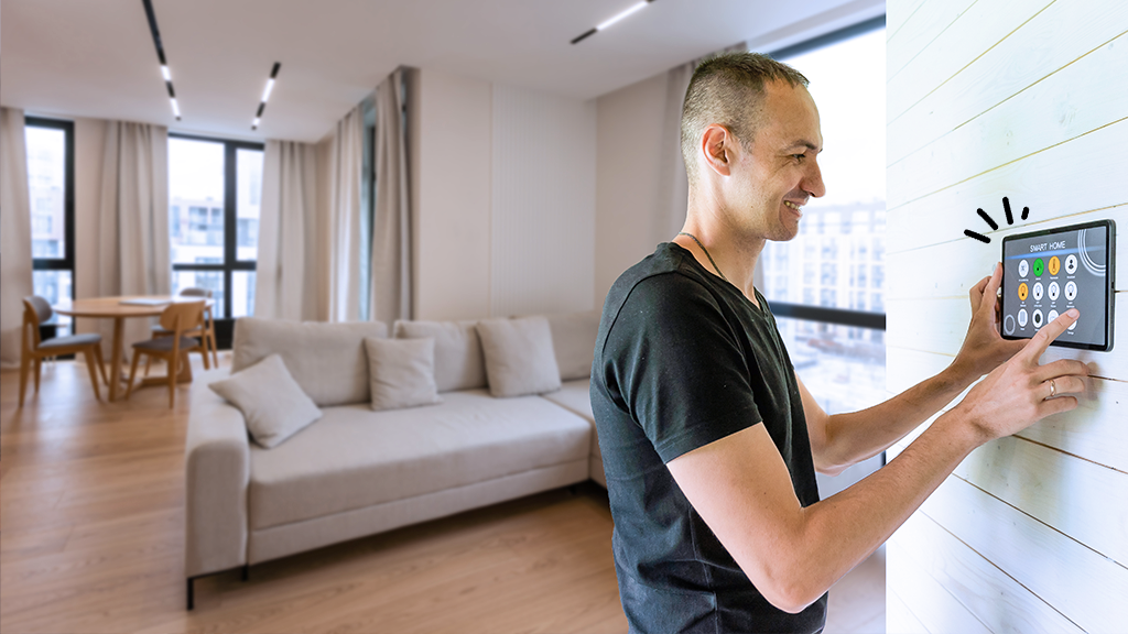 A man using a smartphone mounted on the wall to control smart home devices in a modern living room with large windows, a beige sofa, and a dining area.