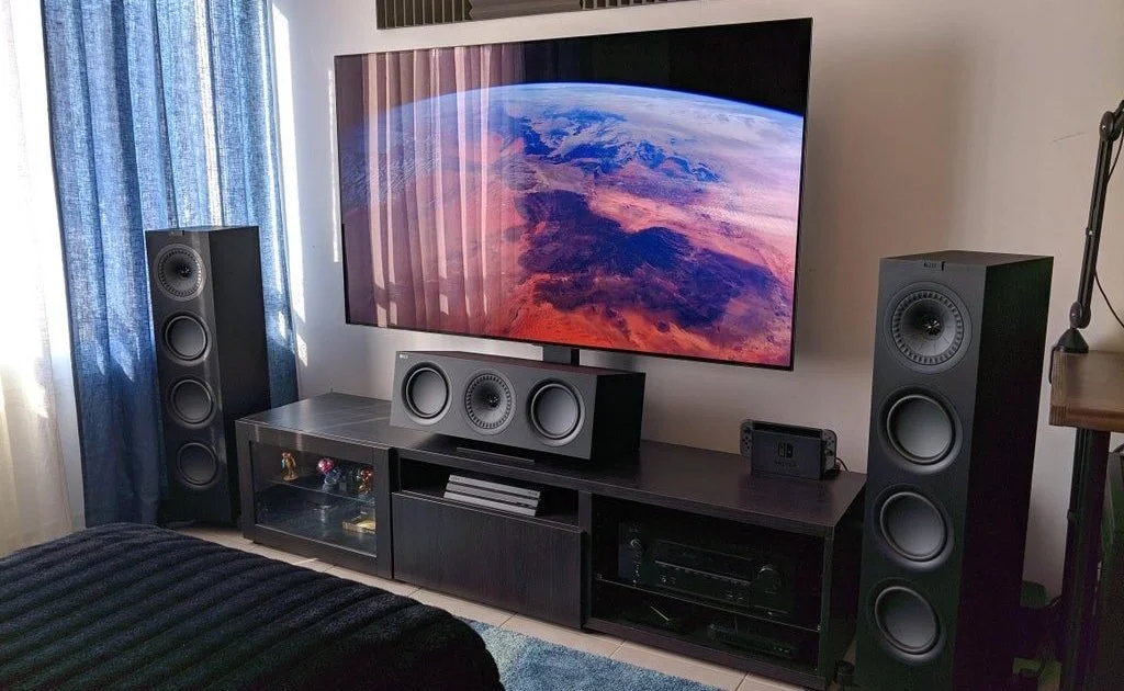 Living room with a large flat-screen TV displaying an image of earth from space, surrounded by tall speakers on each side, a black TV stand with media equipment, and a blue curtain covering a window.