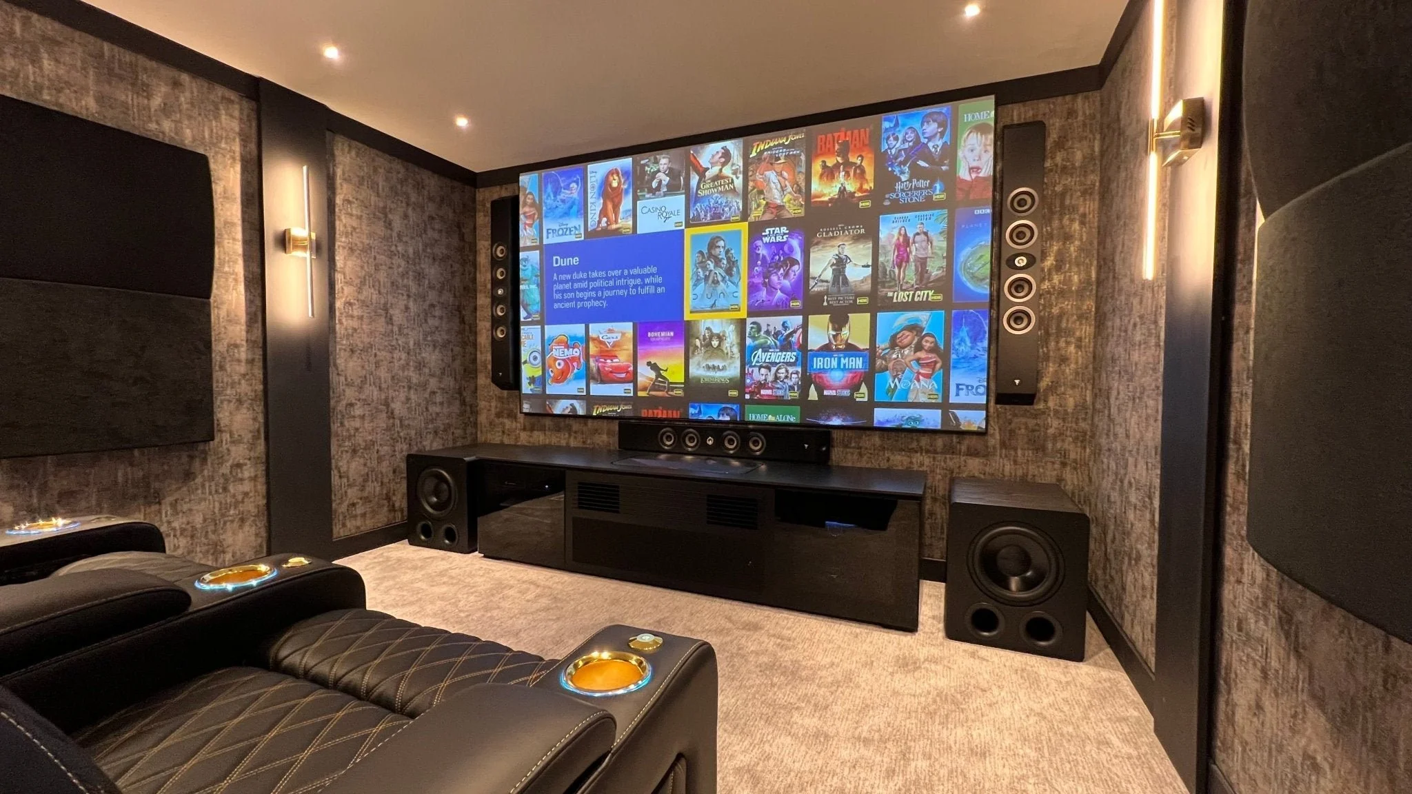 View of a modern home theater with a large screen displaying a streaming service's movie selection, surrounded by black speaker speakers, leather recliner seats with built-in cup holders, and textured walls.