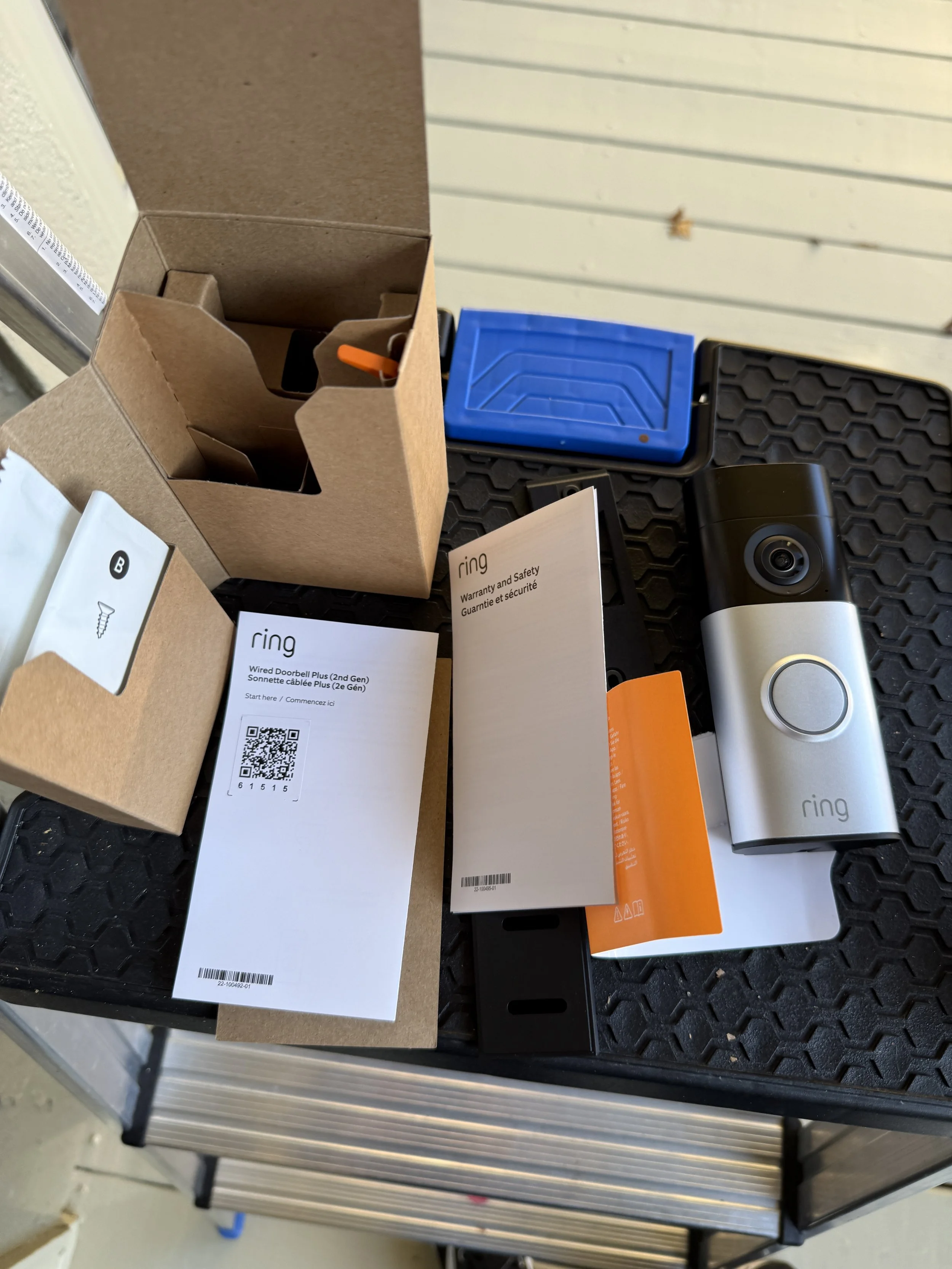 Unboxed Ring doorbell and accessories on a black surface, including instructions, a mounting bracket, a blue storage container, and packaging materials.