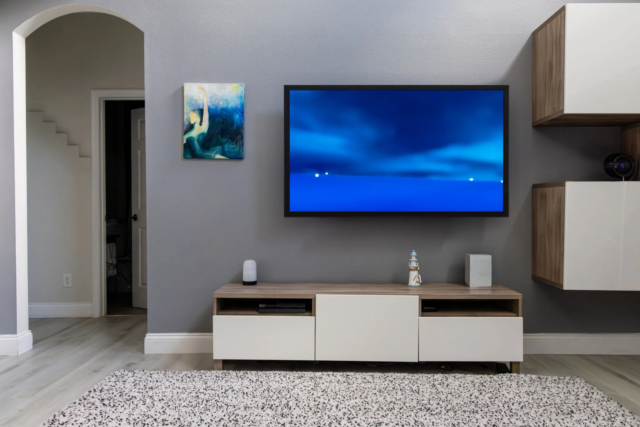 Living room with a wall-mounted flat-screen TV displaying a blue abstract background, a wooden and white entertainment unit with decorative items, and a textured rug on light-colored wood flooring.