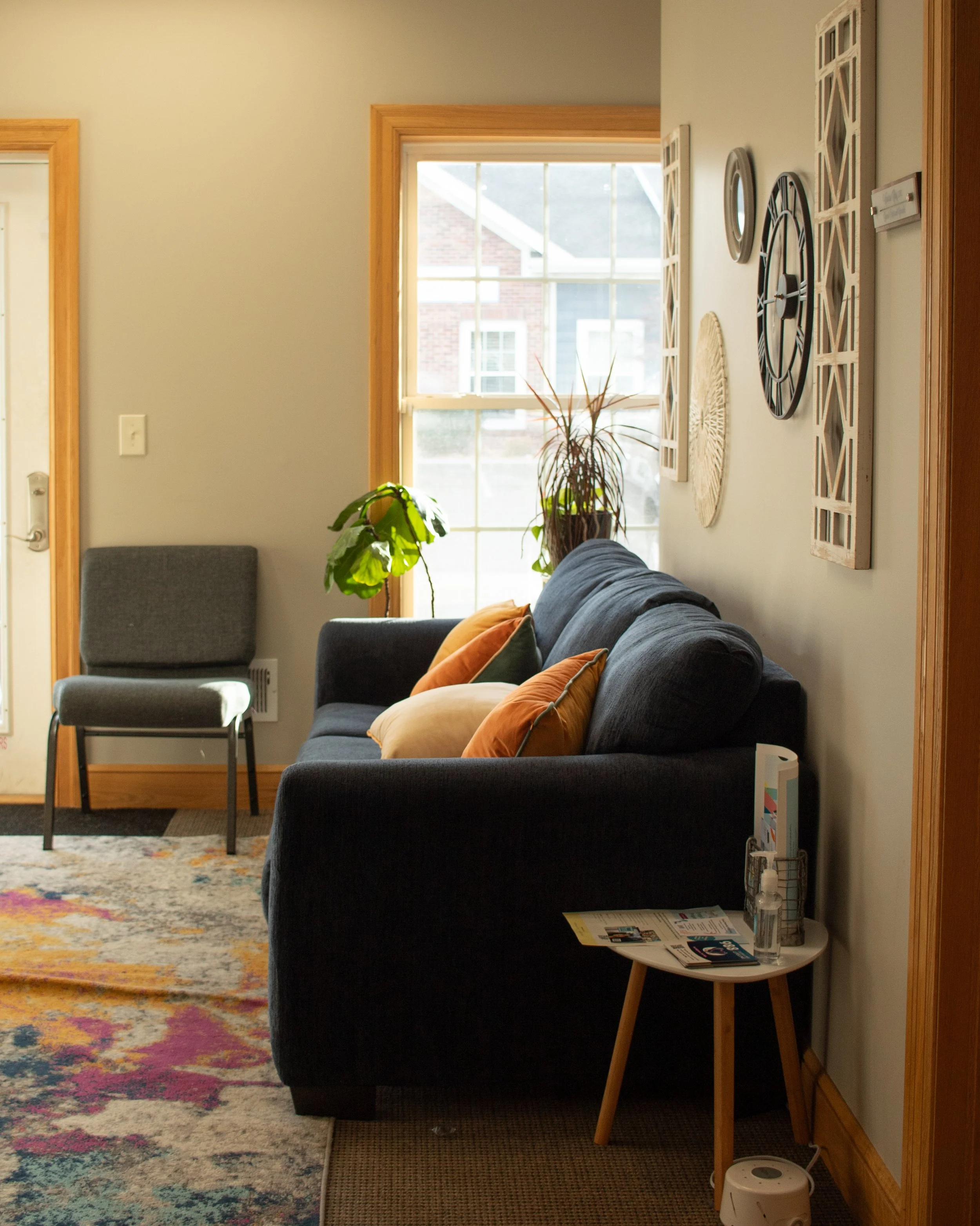 Living room with a dark gray sofa, three throw pillows, a side table with brochures and hand sanitizer, a window letting in natural light, wall art, and a chair near the door.
