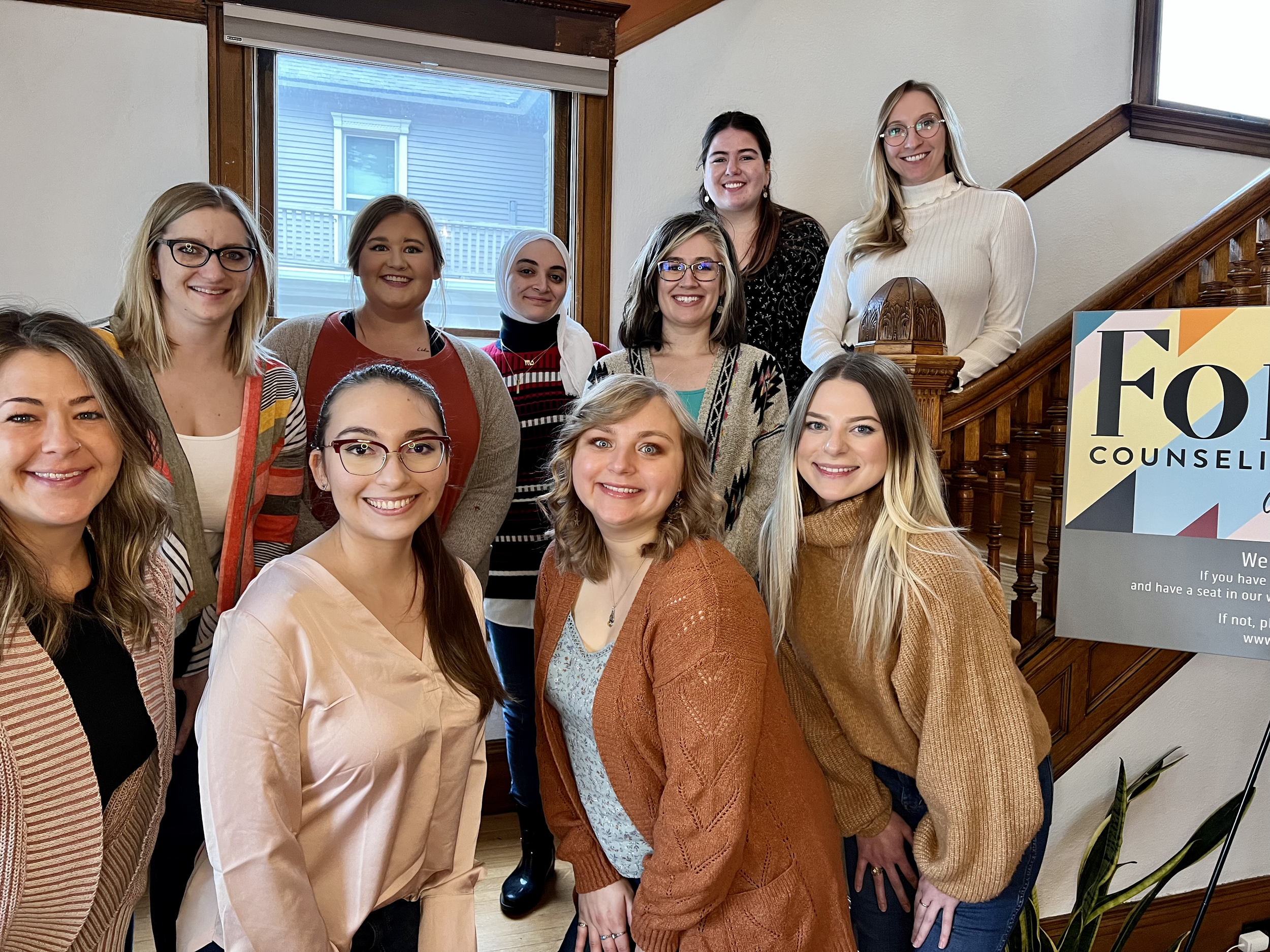 Group of interns smiling and posing for a photo inside a house near a staircase with a sign that reads 'Forge Counseling'.