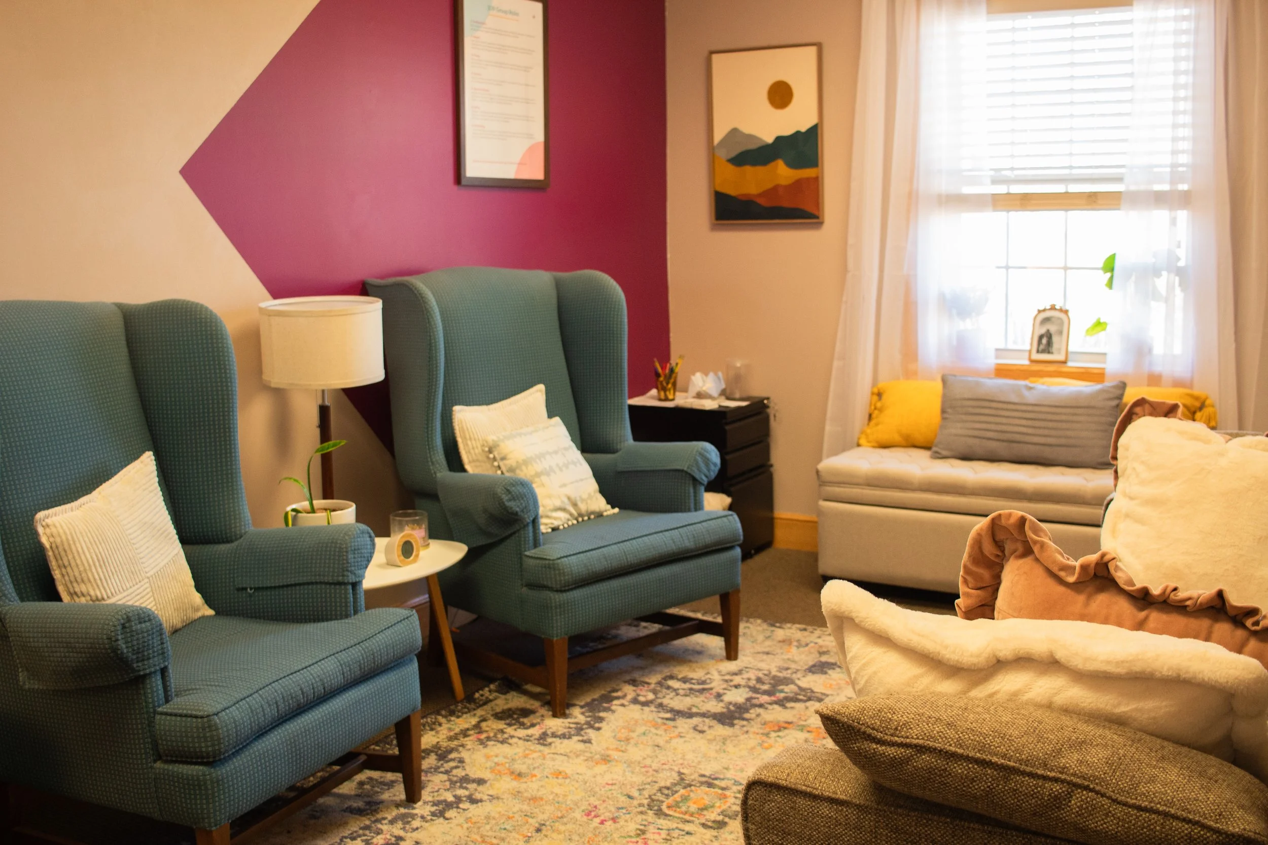 Cozy living room with teal wingback chairs, a side table with a lamp and plant, a window seat with cushions, a patterned rug, and framed art on the wall.