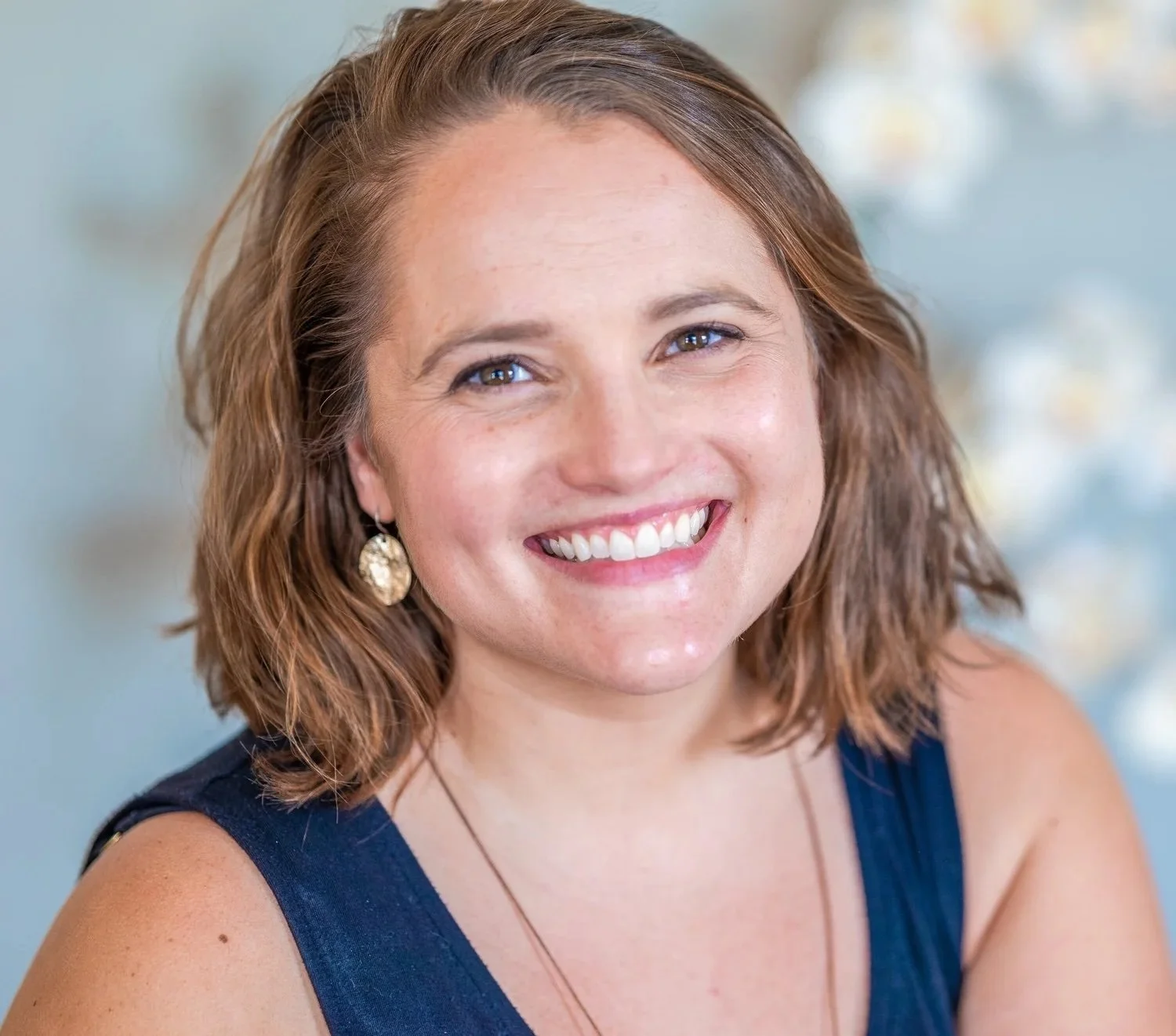 Close-up portrait of a smiling woman with shoulder-length wavy brown hair, wearing earrings and a dark top, with a light blurred background.
