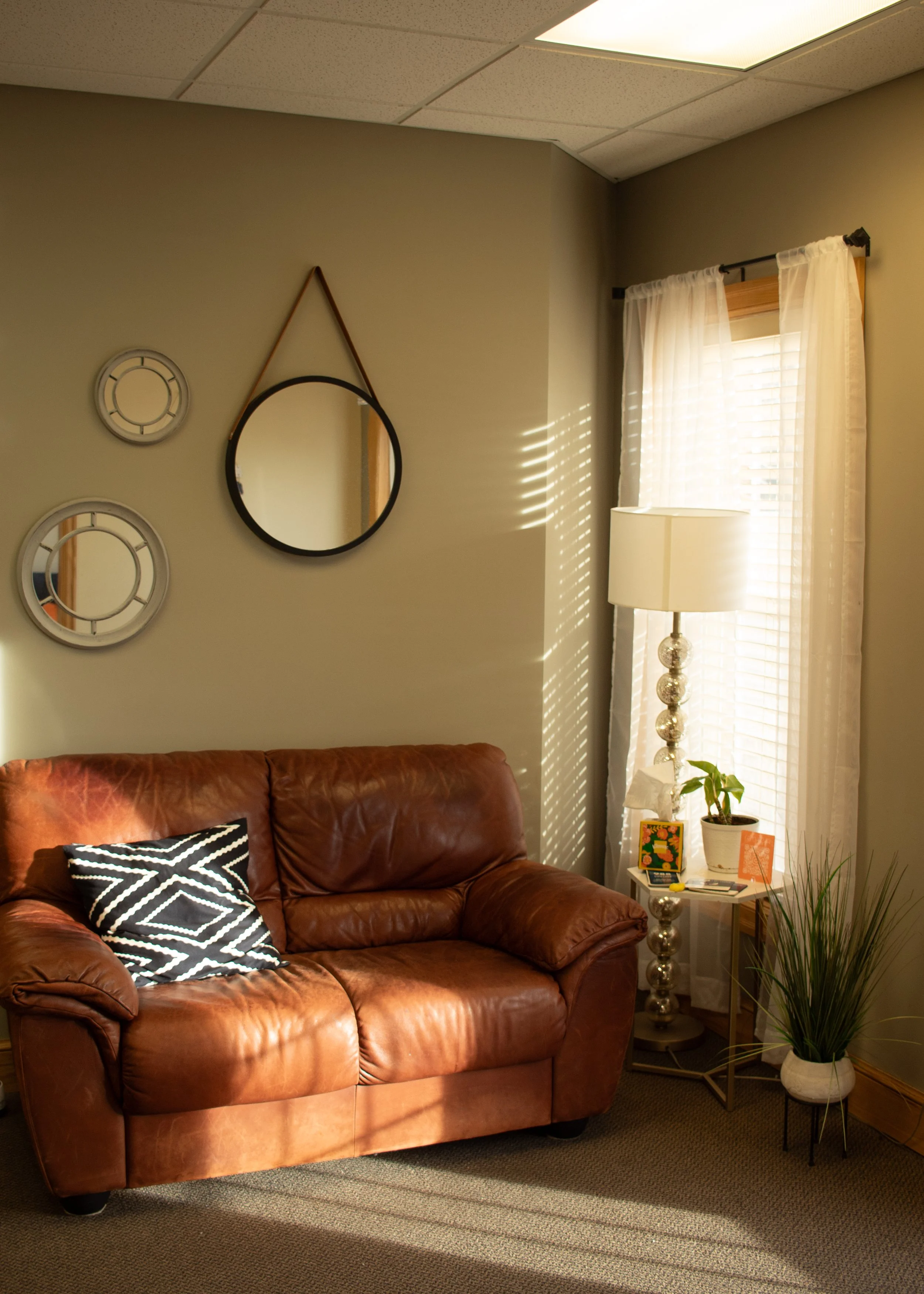 A cozy living room corner with a brown leather sofa, a black and white patterned pillow, a small side table with a potted plant, tissue box, and framed picture, a tall floor lamp with decorative glass balls, a window with sheer white curtains, wall mirrors, and a wall clock, illuminated by sunlight streaming through blinds.