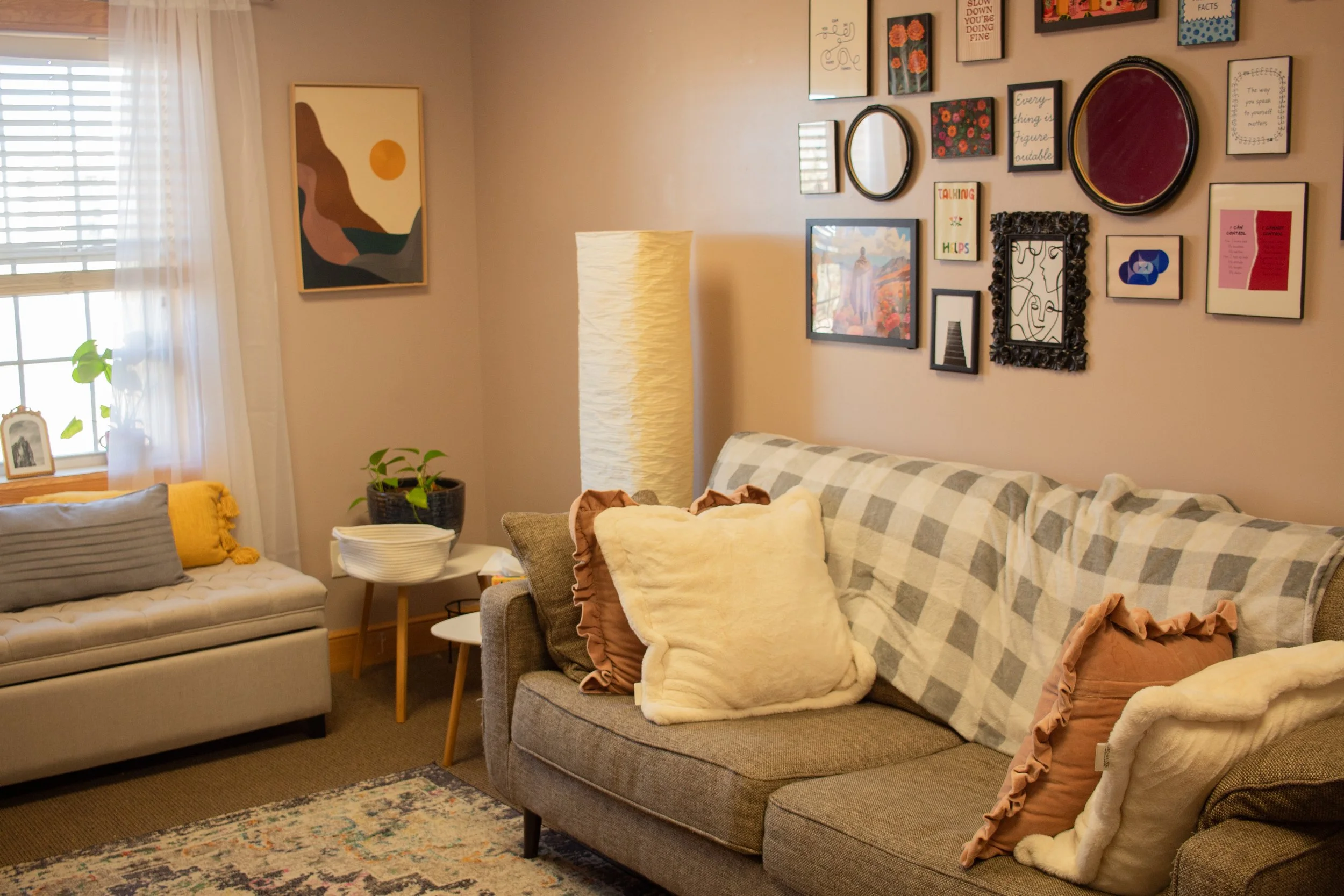 Living room with a gray sofa adorned with plush cushions, a wall gallery of framed art, a tall white floor lamp, a window with sheer curtains, and a small table with a potted plant and decorative items.