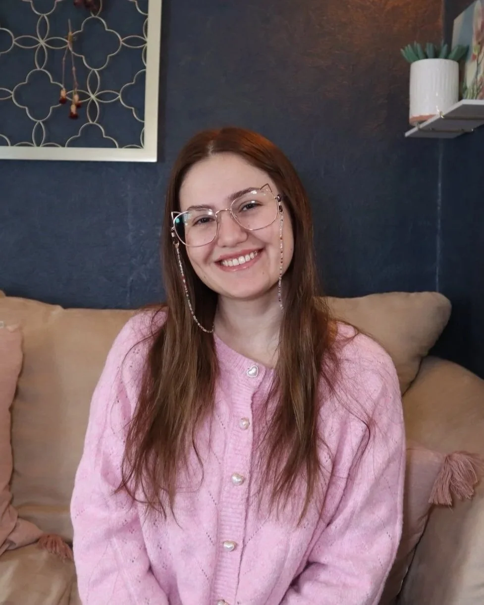 A young woman with long, wavy brown hair and glasses smiling at the camera while sitting on a beige sofa in a cozy living room, wearing a pink cardigan.