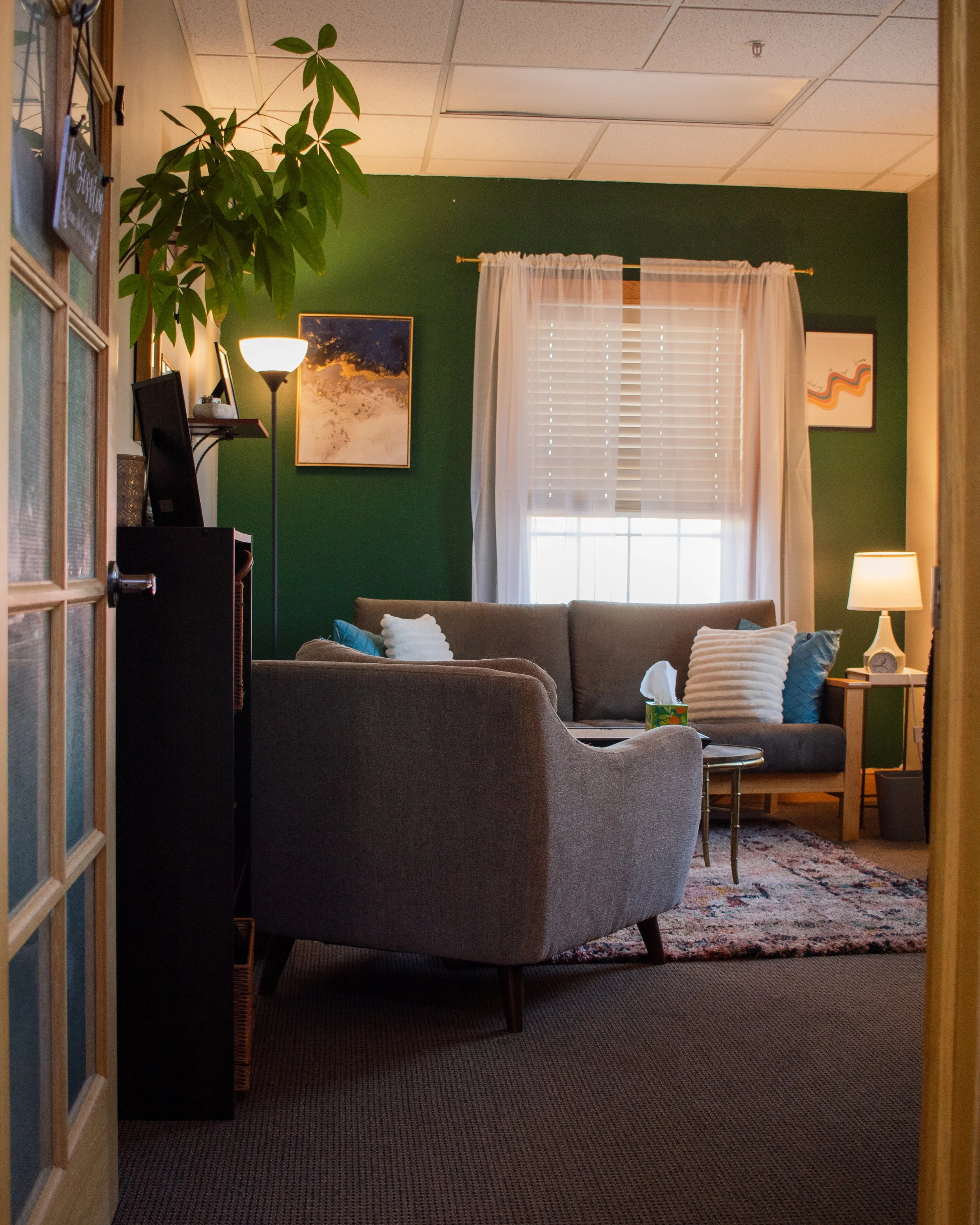 A cozy living room with green walls, a large window with white curtains, a gray sofa with white and blue pillows, a small wooden side table with a lamp, a patterned rug, and artwork on the walls. There is also a tall potted plant and a floor lamp near the sofa.