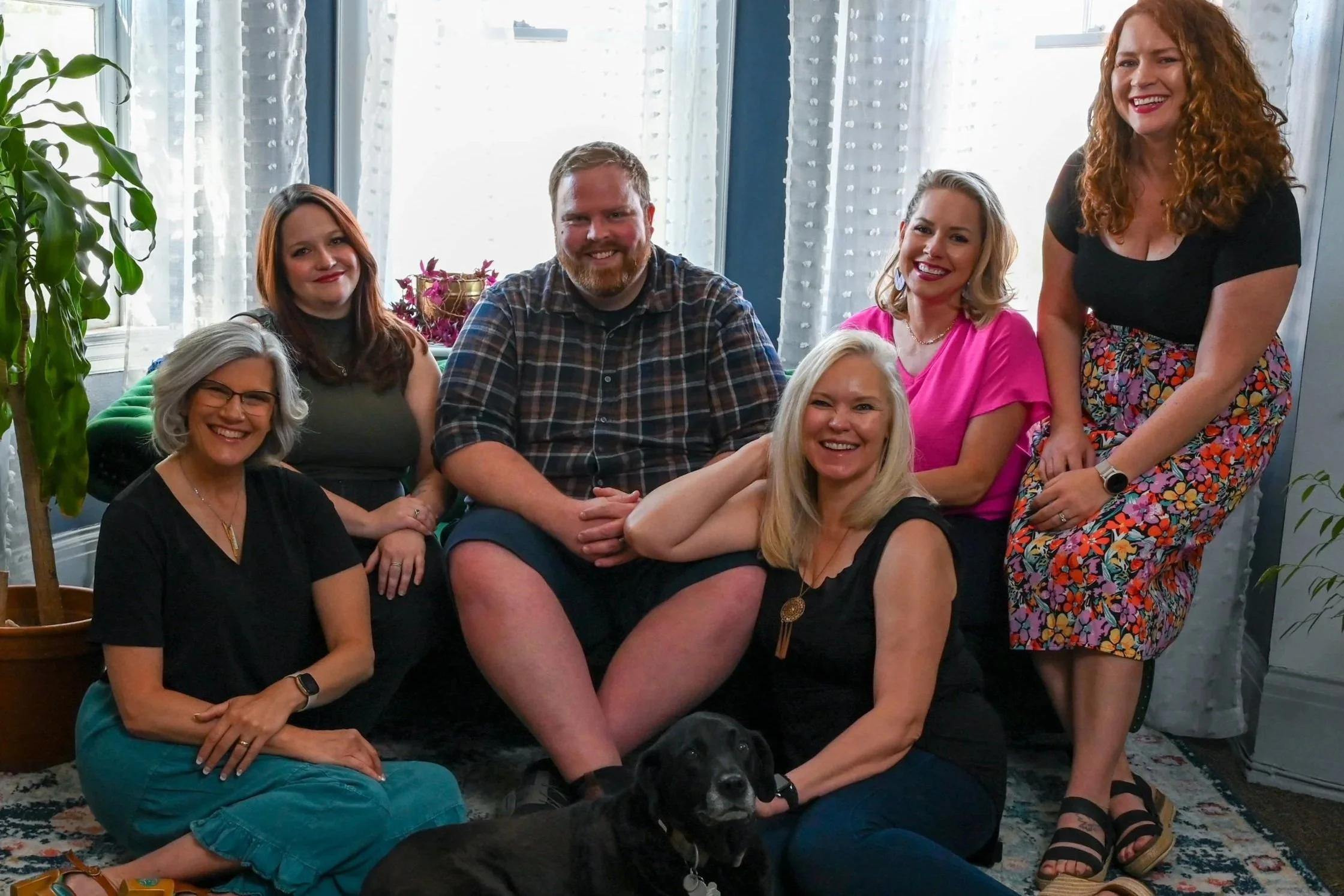 Group of seven therapists, posing together in a cozy room with light coming through curtains. Some are sitting, some standing, with a dog lying on the floor in front.
