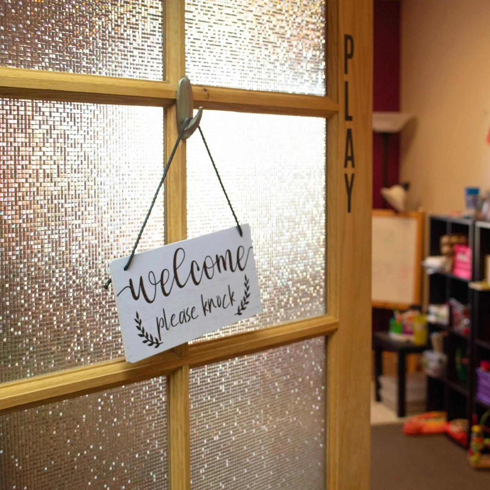 A wooden door with a glass panel and a sign that says "welcome please knock" hanging from a nail in the door. The sign has decorative elements and is positioned near the door handle.
