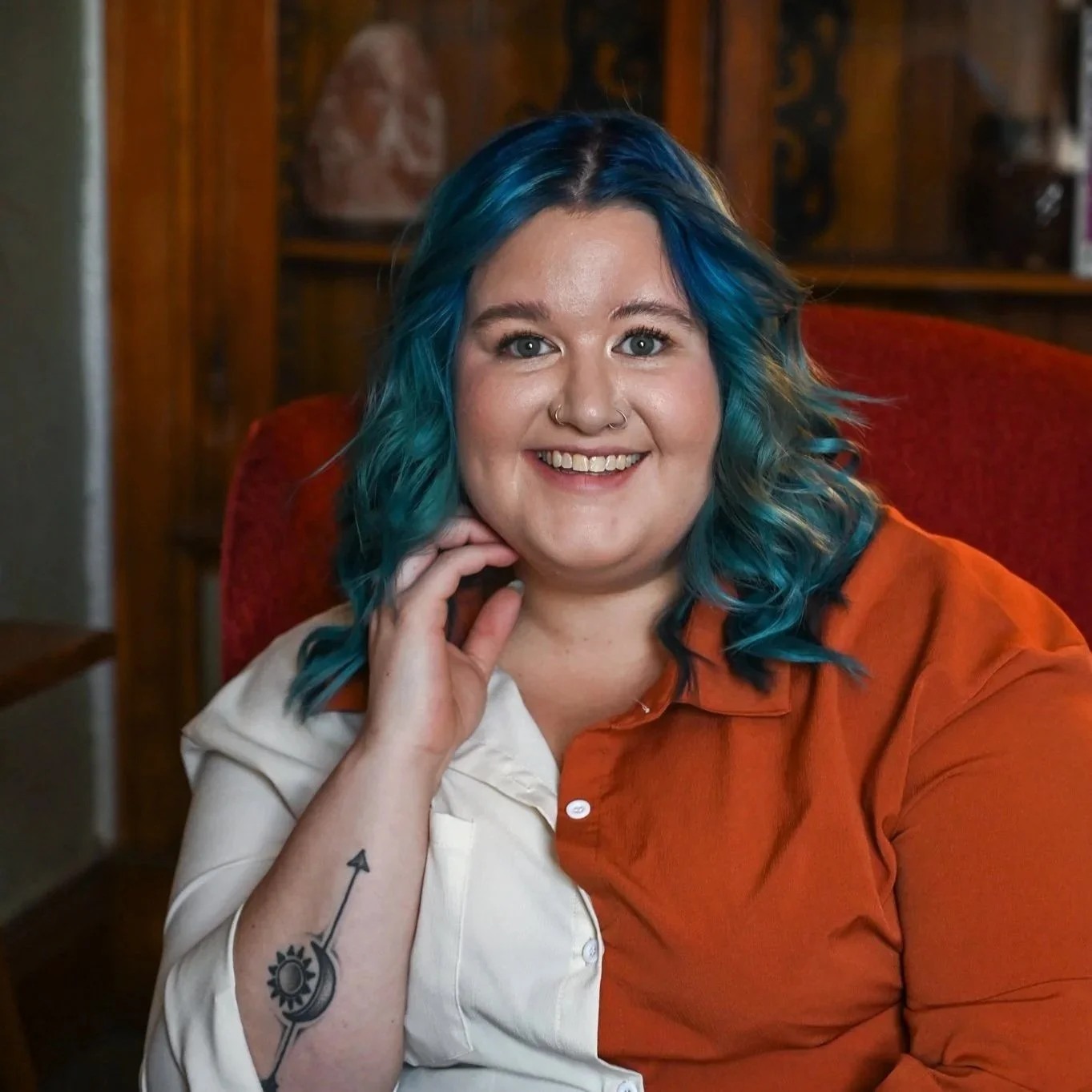 A woman with multicolored blue hair smiling, sitting in a red armchair with wooden shelves and decorative items in the background.