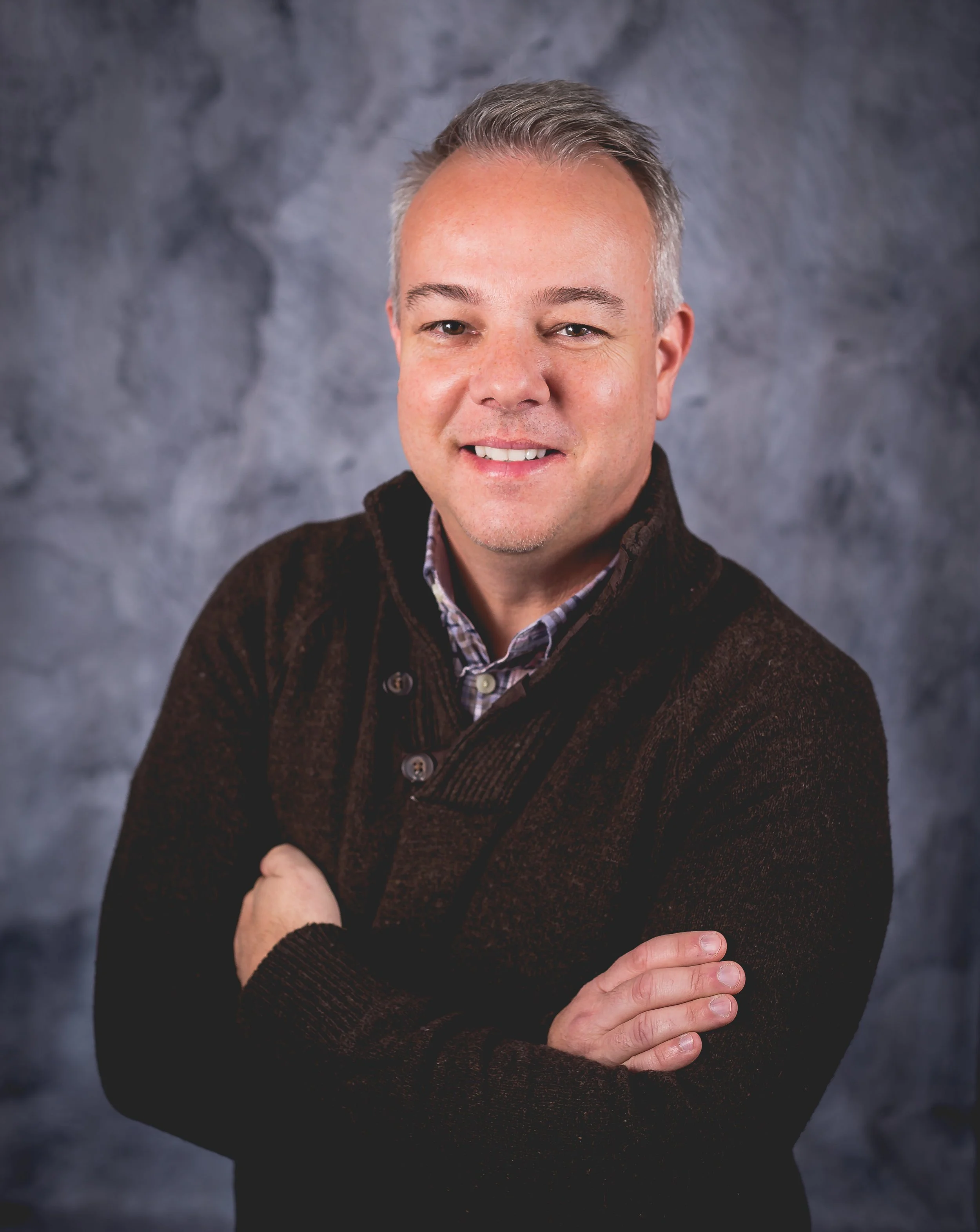 A middle-aged man with short gray hair smiling, wearing a dark brown corduroy sweater over a collared shirt, standing with his arms crossed in front of a gray textured background.