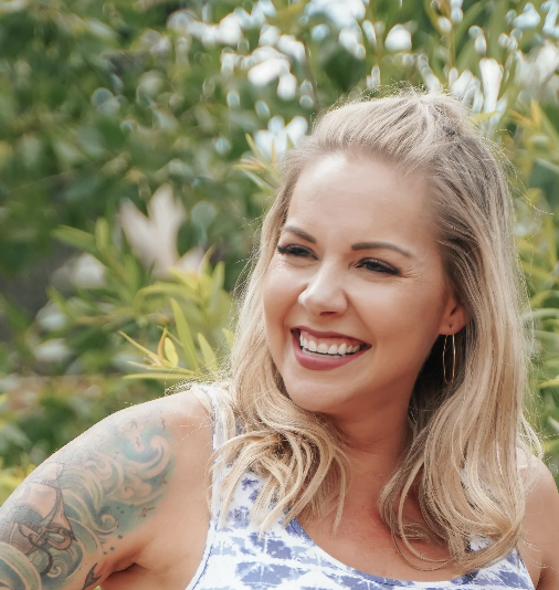 Young woman with long blonde hair smiling outdoors, wearing a sleeveless top with a tattoo on her arm, and hoop earrings, surrounded by green foliage.