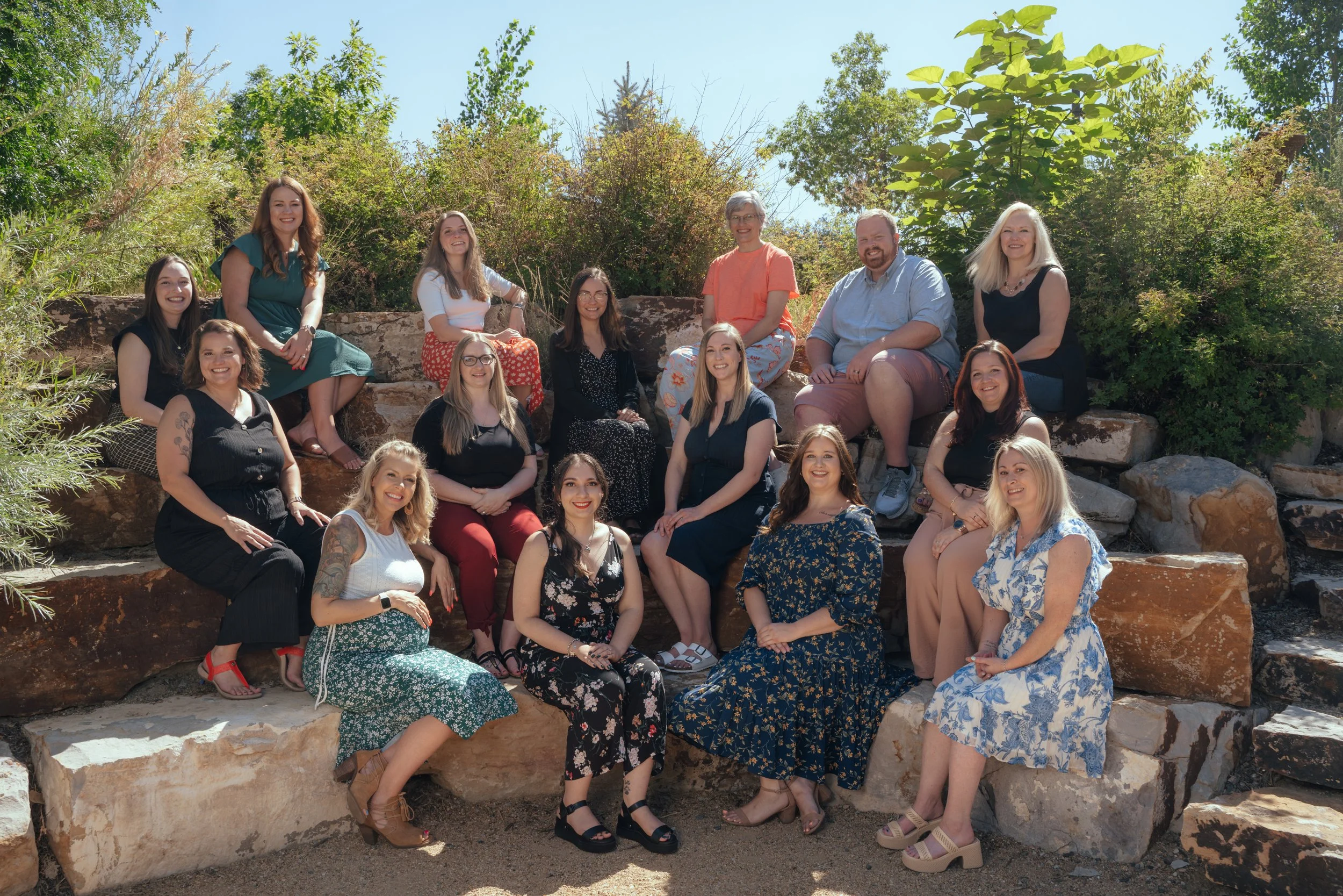 Group of 16 women and 2 men sitting on rocks outdoors with greenery in the background, smiling at the camera