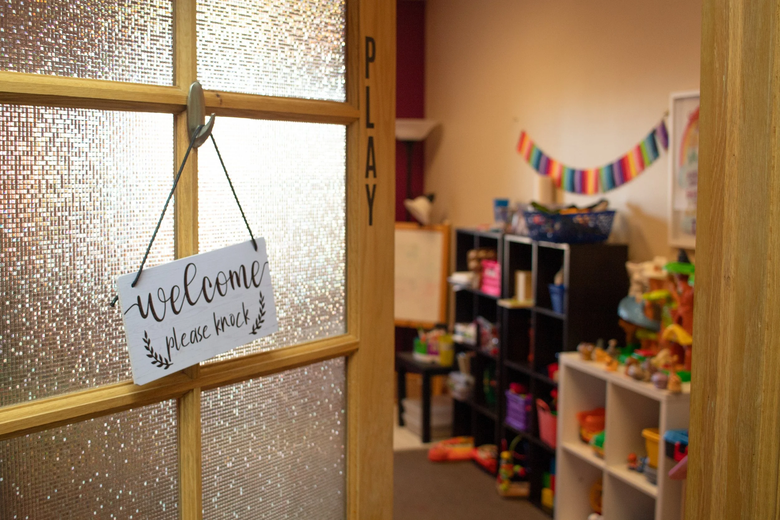 A door with a sign that says "welcome please knock" opening into a play therapy room with shelves filled with toys and supplies, a colorful paper chain decoration on the wall, and a small table with more toys.