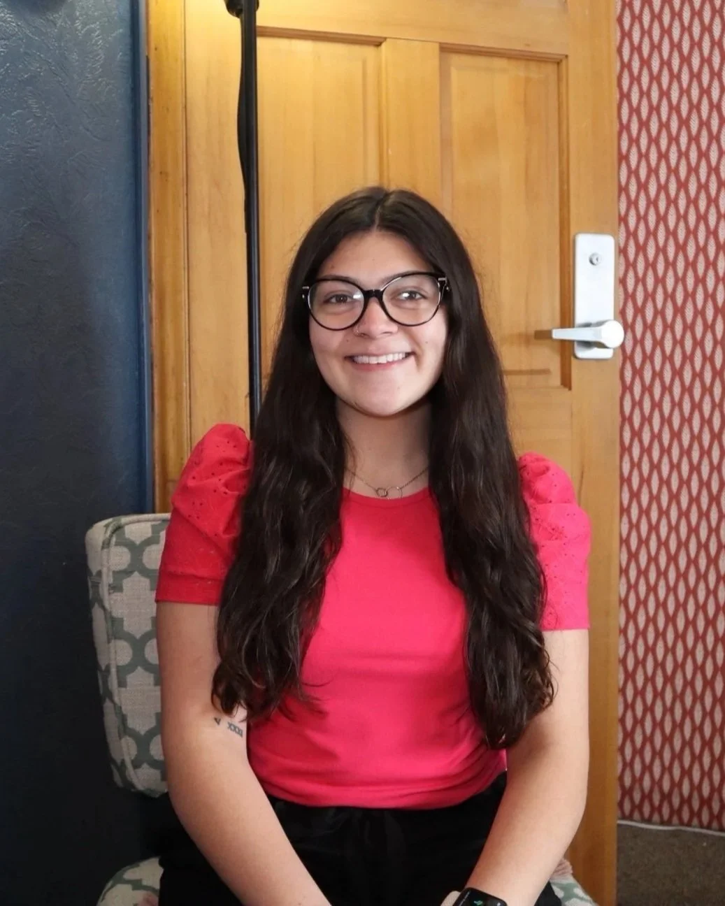 A young woman with long dark hair, glasses, and a bright red shirt, smiling and sitting on a patterned chair, with a wooden door and patterned wallpaper in the background.
