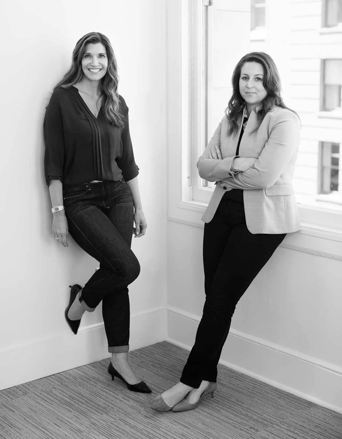 Black and white photo of two women in business casual attire posing in a corner near a window with city buildings visible outside. One woman is leaning against the wall, smiling with her hands at her sides, and the other is standing with arms crossed, looking at the camera.