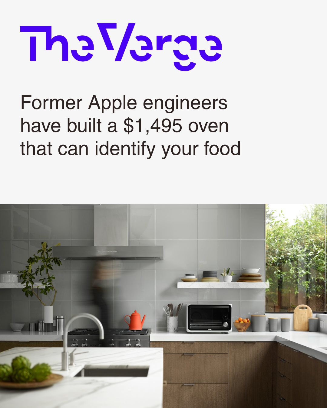 The Verge logo with headline about a $1,495 oven that can identify food, over a modern kitchen with gray tiled walls, a stove with a red teapot, white marble countertops, and a window with greenery outside.