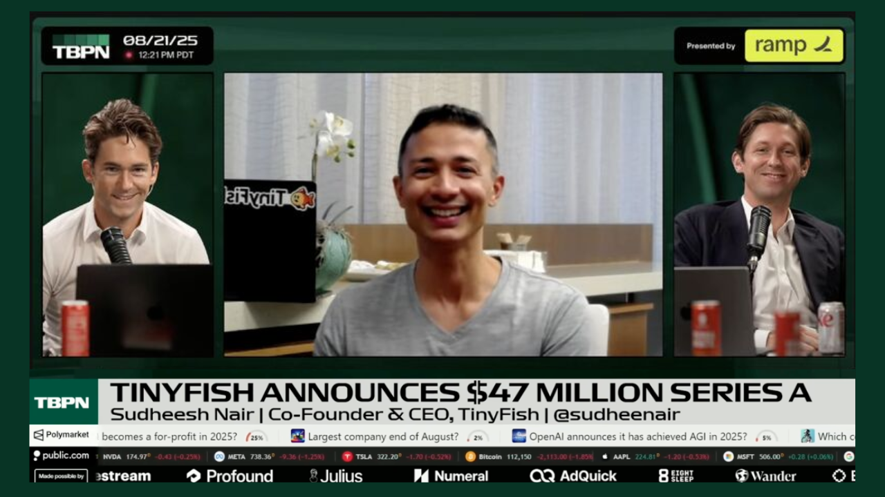 A televised news segment features three men with headsets, microphones, and laptop computers, discussing a financial announcement. The central person is smiling in front of a neutral background, while the two men on either side are also smiling, sitt