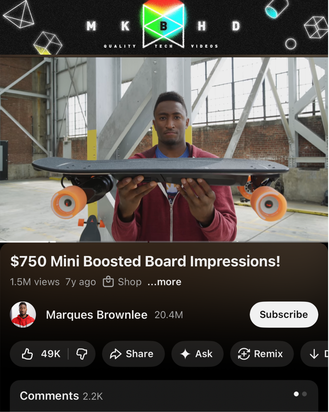 Screenshot of a video review by YouTuber Marques Brownlee. He's holding a Boosted Board skateboard inside a large warehouse. The skateboard has orange wheels.
