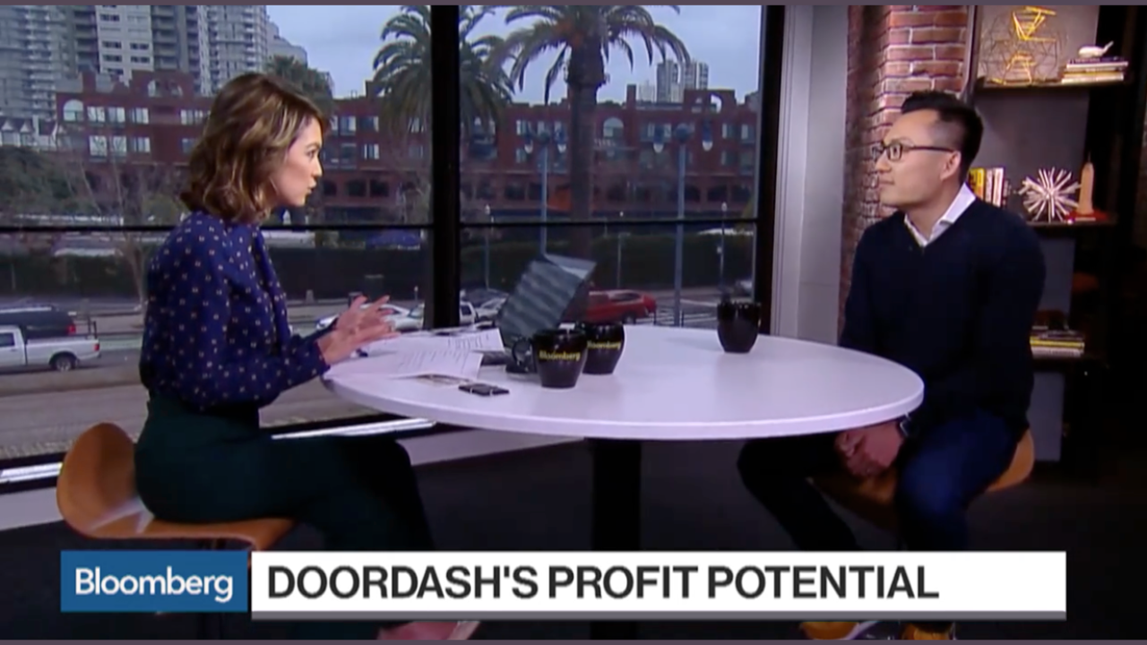 News interview at a table with two people, a woman and a man, with a cityscape and palm trees visible through large windows behind them. The woman is speaking, with papers and a laptop on the table. The man listens attentively. The news banner reads 