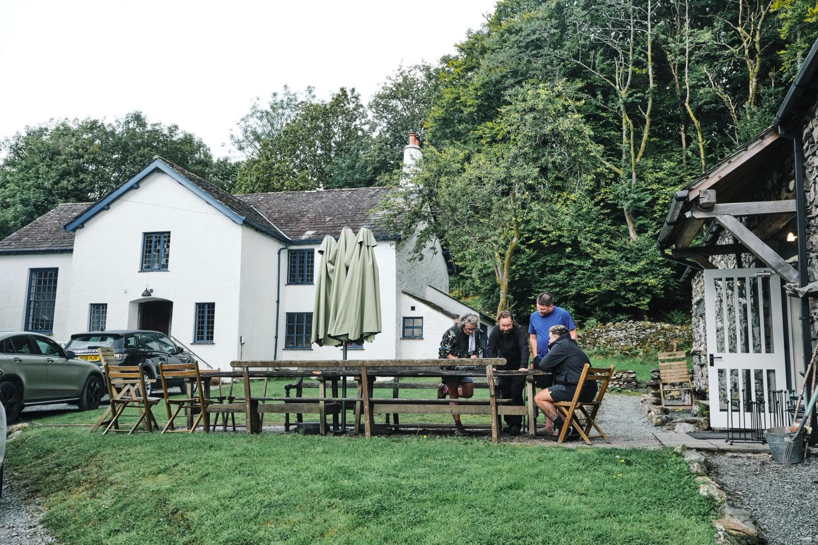 five people gathered around a wooden outdoor table on a patio in front of a white house with black framed windows, surrounded by cars and greenery.