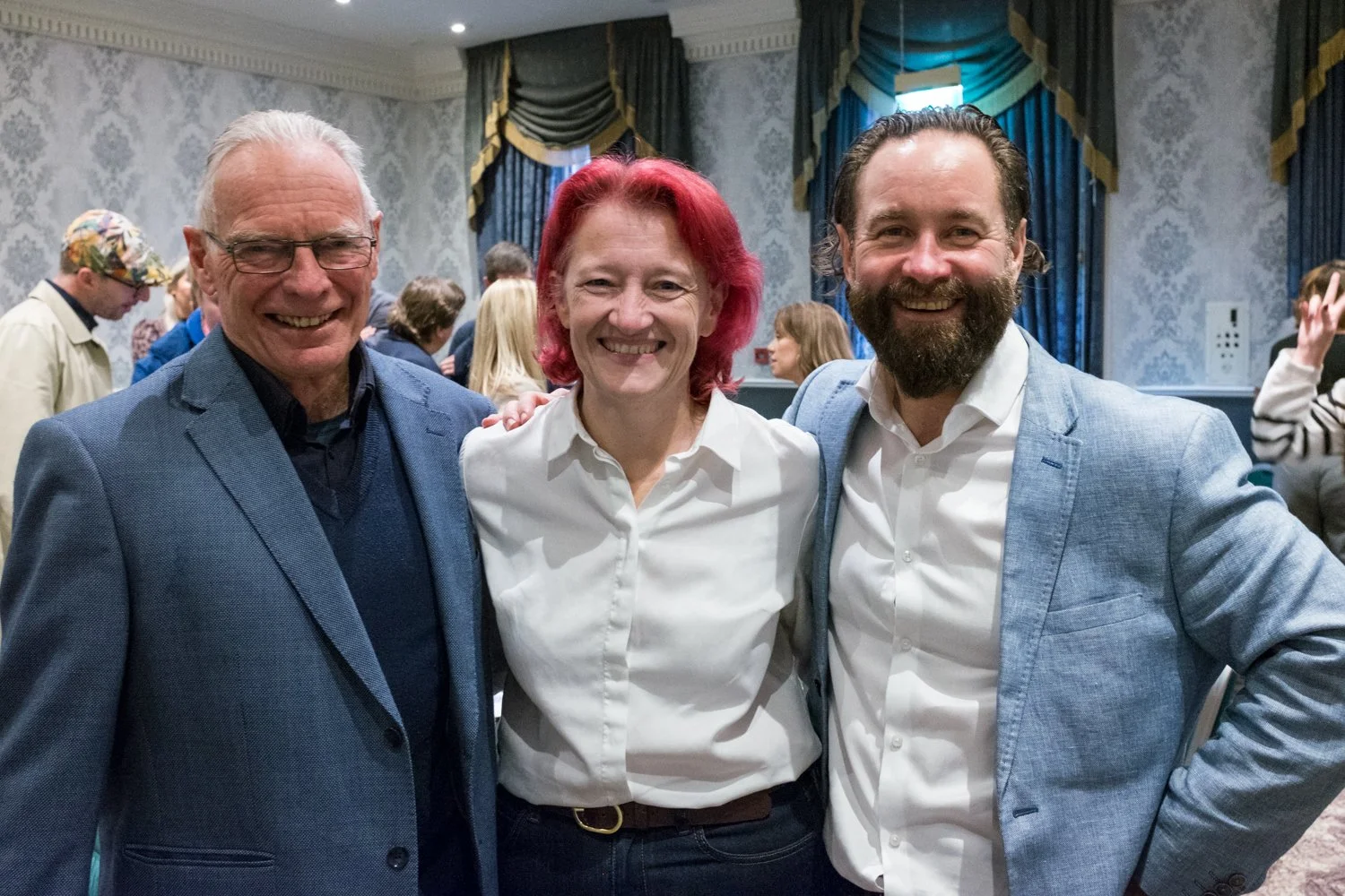 Three smiling people standing close together indoors at a social event, with a crowd in the background. The person on the left is an older man with white hair, glasses, wearing a blue blazer. The person in the middle is a woman with short red hair, wearing a white shirt. The person on the right is a man with dark hair and a beard, wearing a light blue blazer and white shirt.
