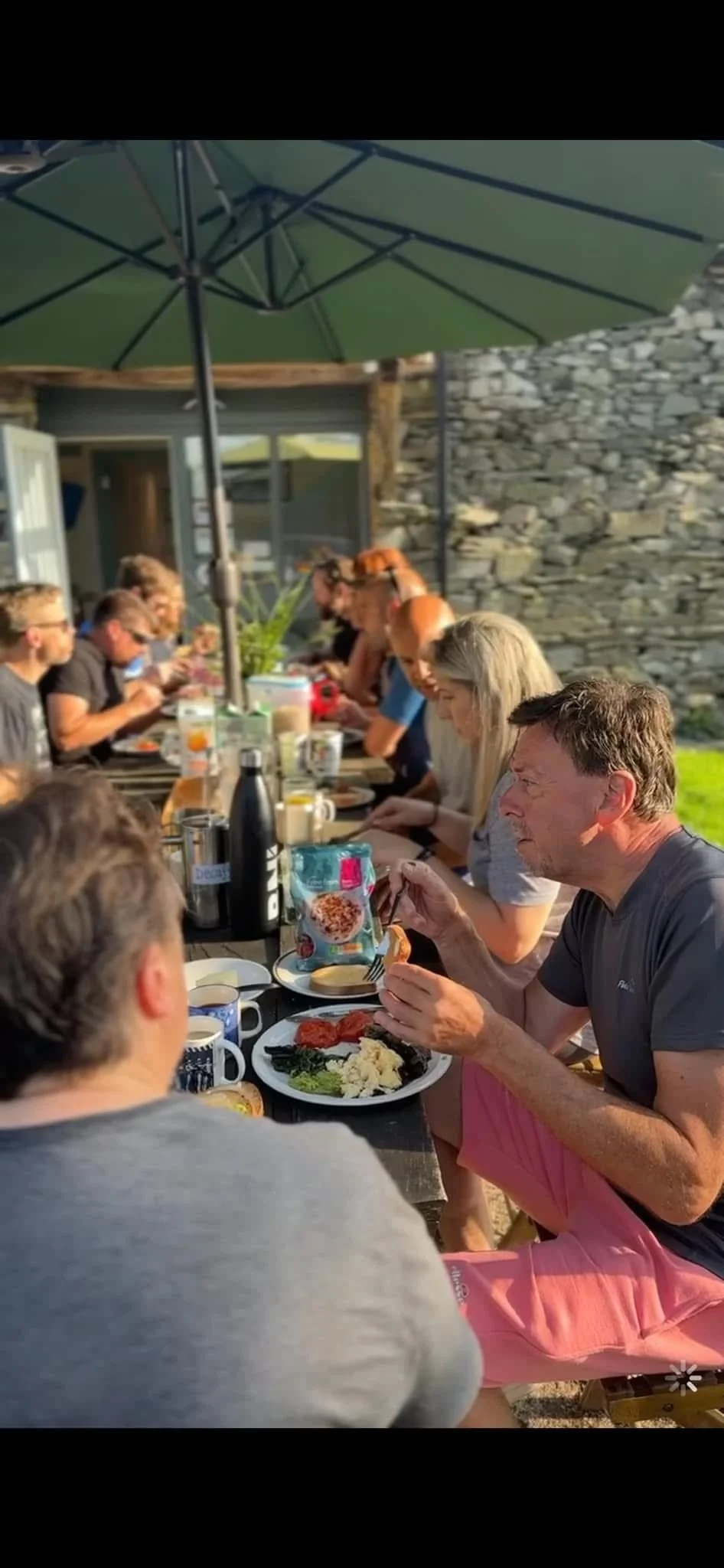 People having a meal outdoors at a long table under a large green umbrella, with a stone wall and a building in the background.