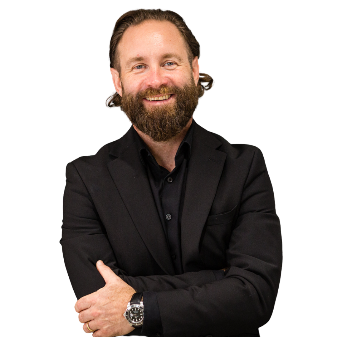 A man with a beard and long hair smiling, wearing a black suit and black shirt, with arms crossed, against a black background.