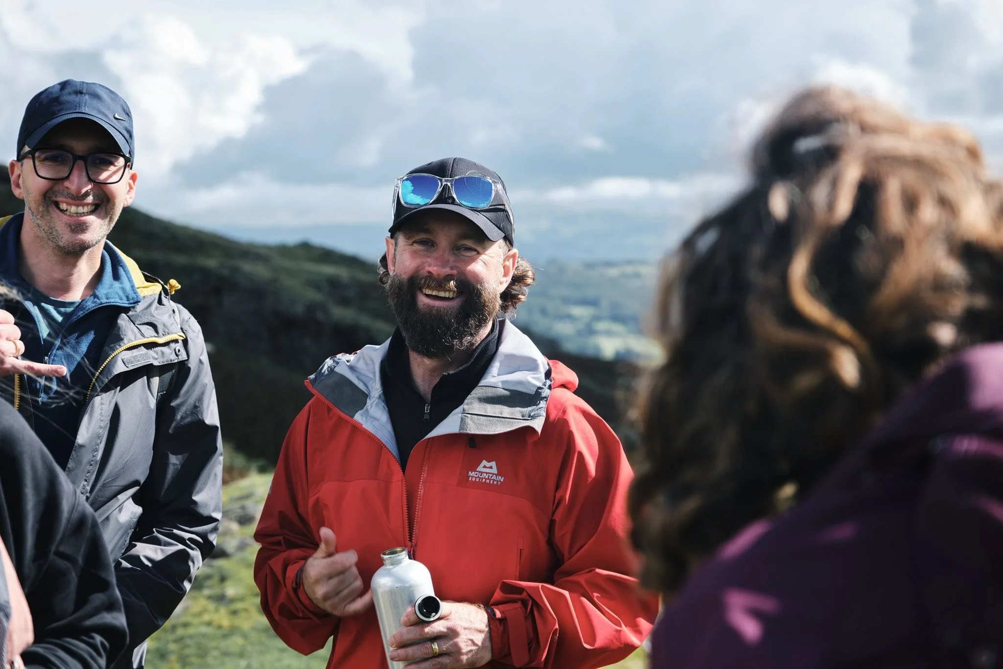 Three people outdoors, smiling and talking, in a mountainous area with scenic views and cloudy sky.