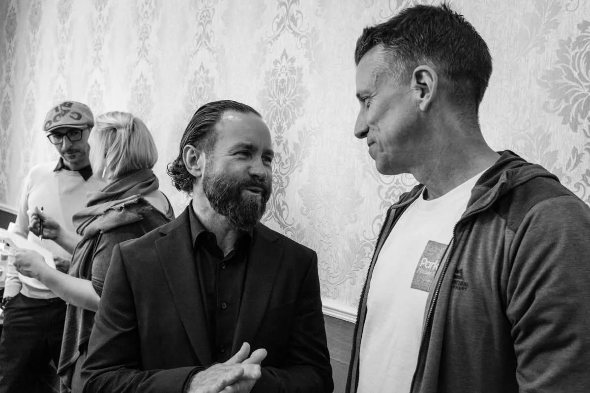 Two men having a conversation, one with a beard in a suit, the other in a casual hoodie, with two people in the background, one taking notes.