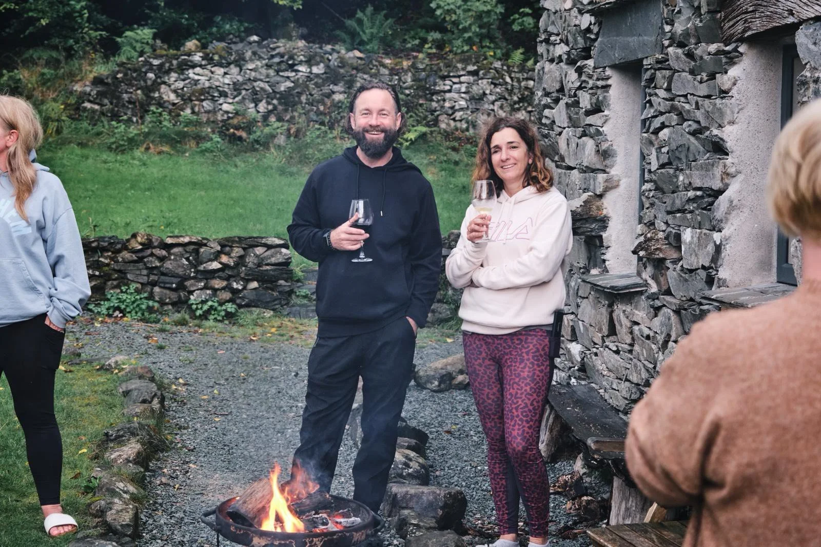 People standing around a campfire, holding wine glasses, outside a stone building in a backyard with green grass and rocks.