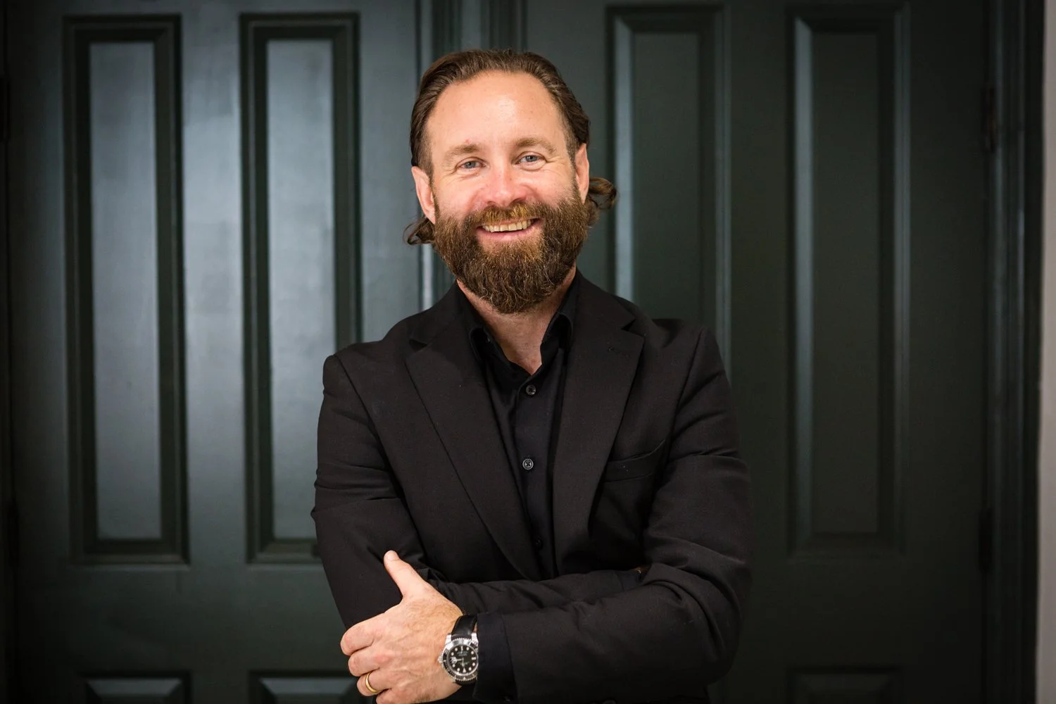 A man with a beard and mustache, wearing a black suit and black shirt, smiling with arms crossed, standing in front of a dark green paneled door.